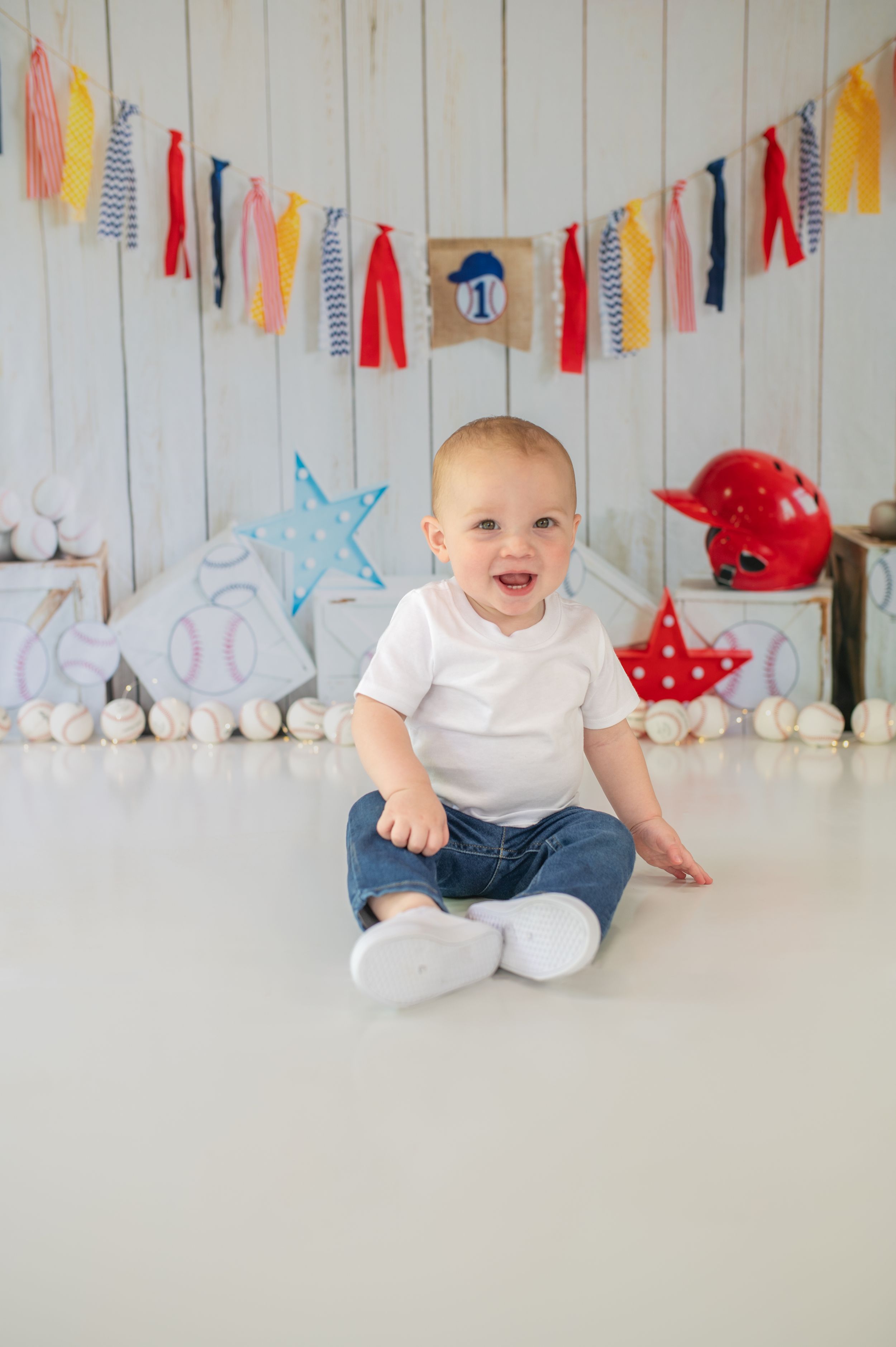 Baby in white shirt and jeans sits among festive party decorations.
