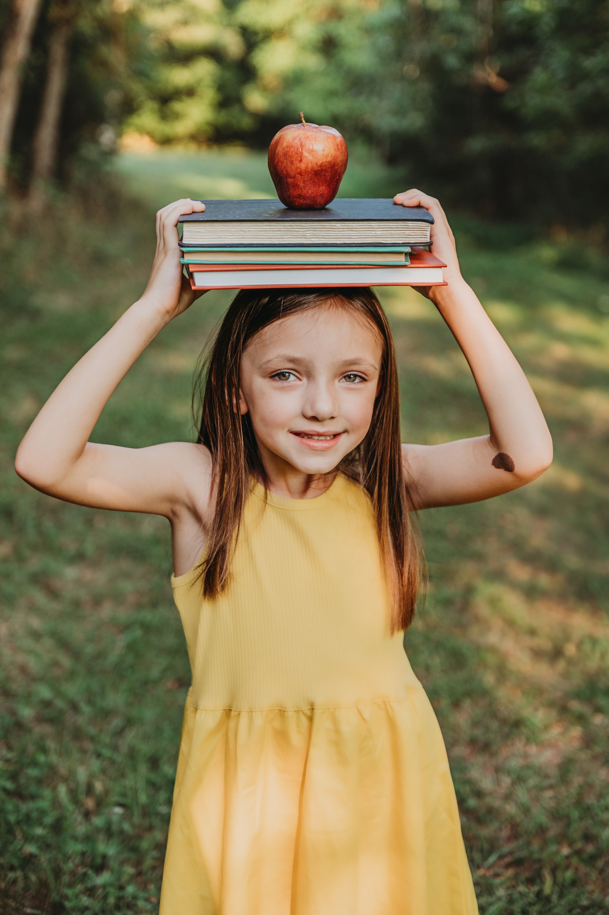 Child in yellow dress balancing books and apple on head in outdoor setting.