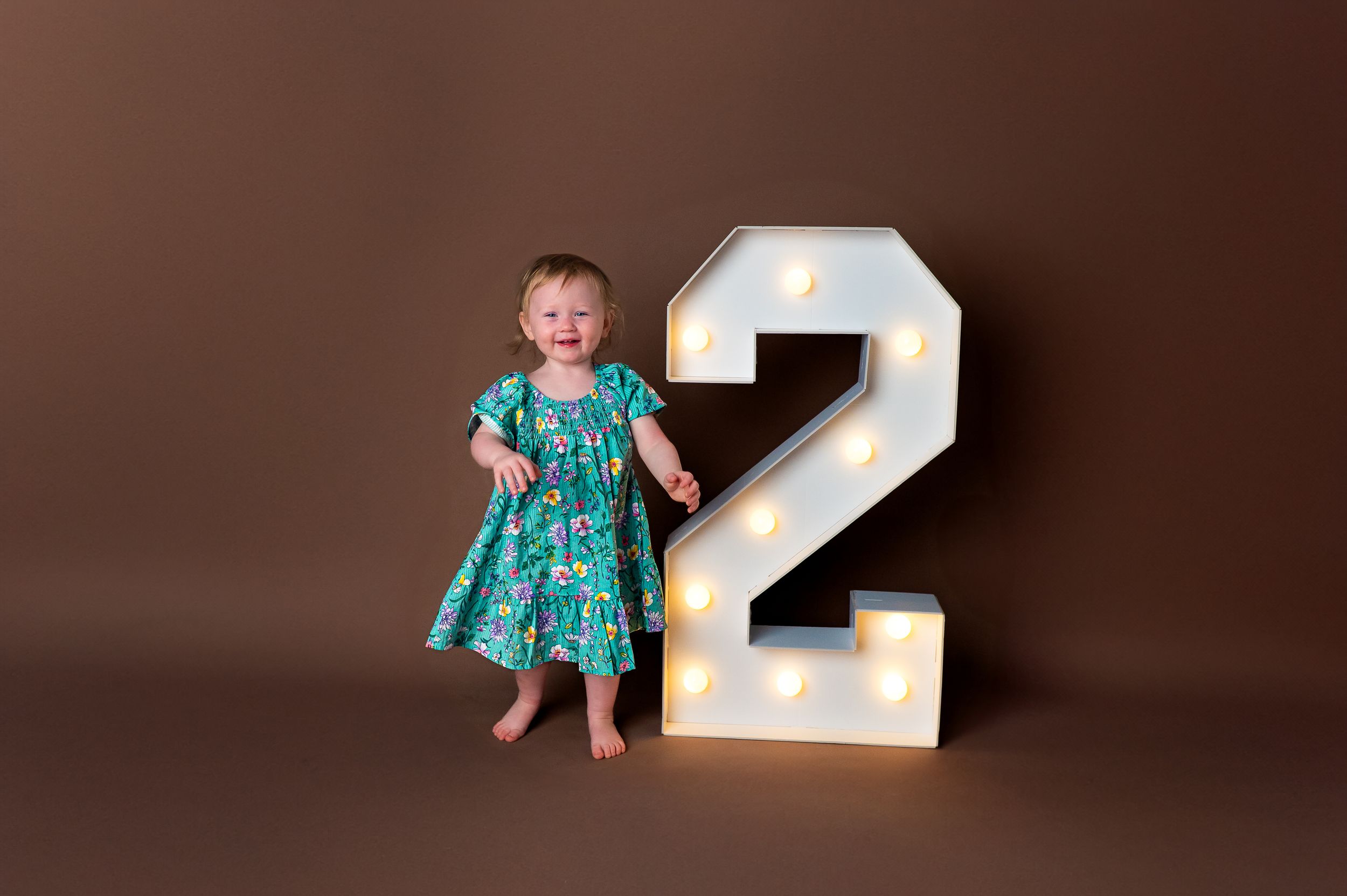 Toddler girl in teal floral dress smiling beside a large illuminated number 2 sign on brown background.