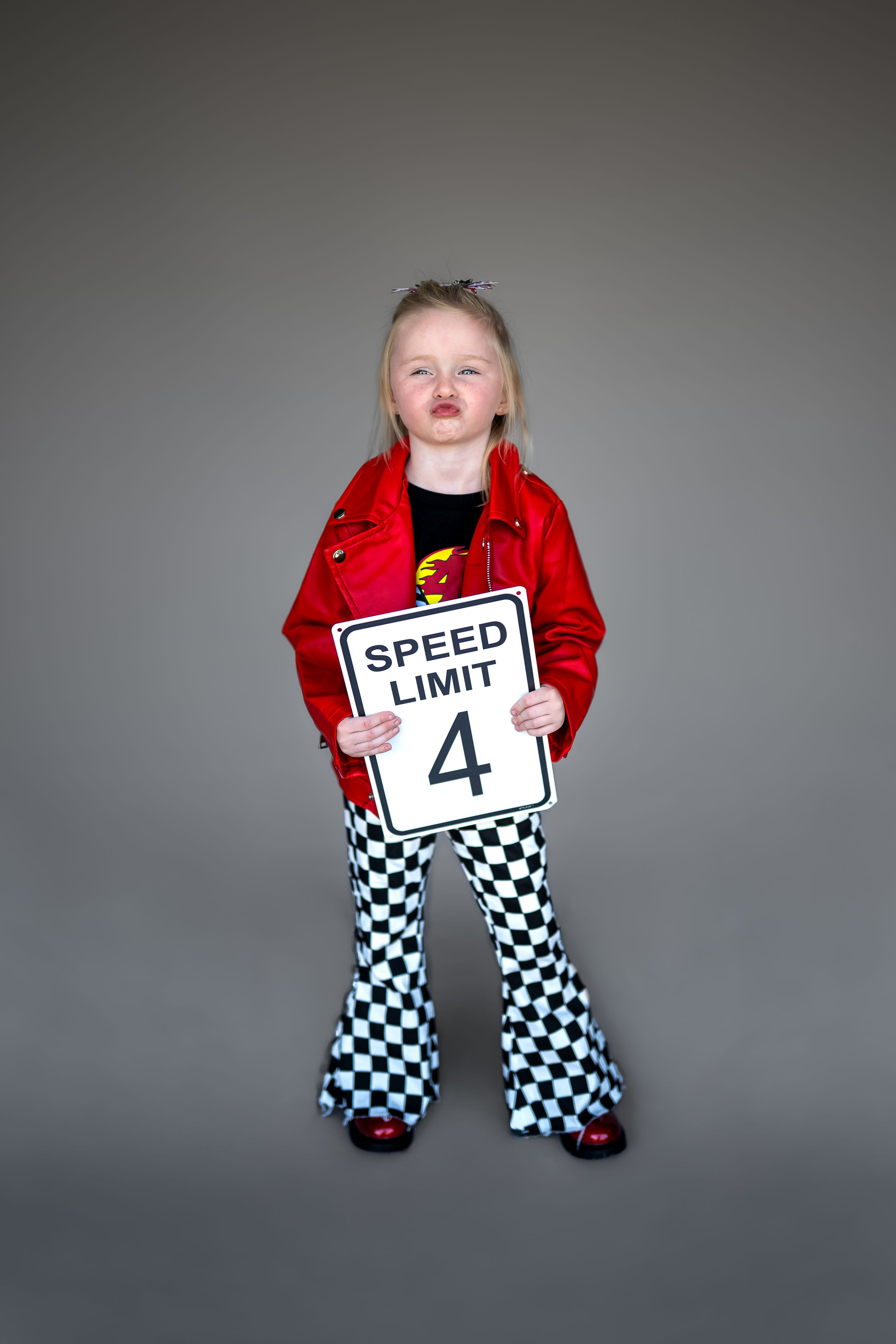 A young girl in a red jacket and checkered pants holds a Speed Limit 4 sign, celebrating her 4th birthday.