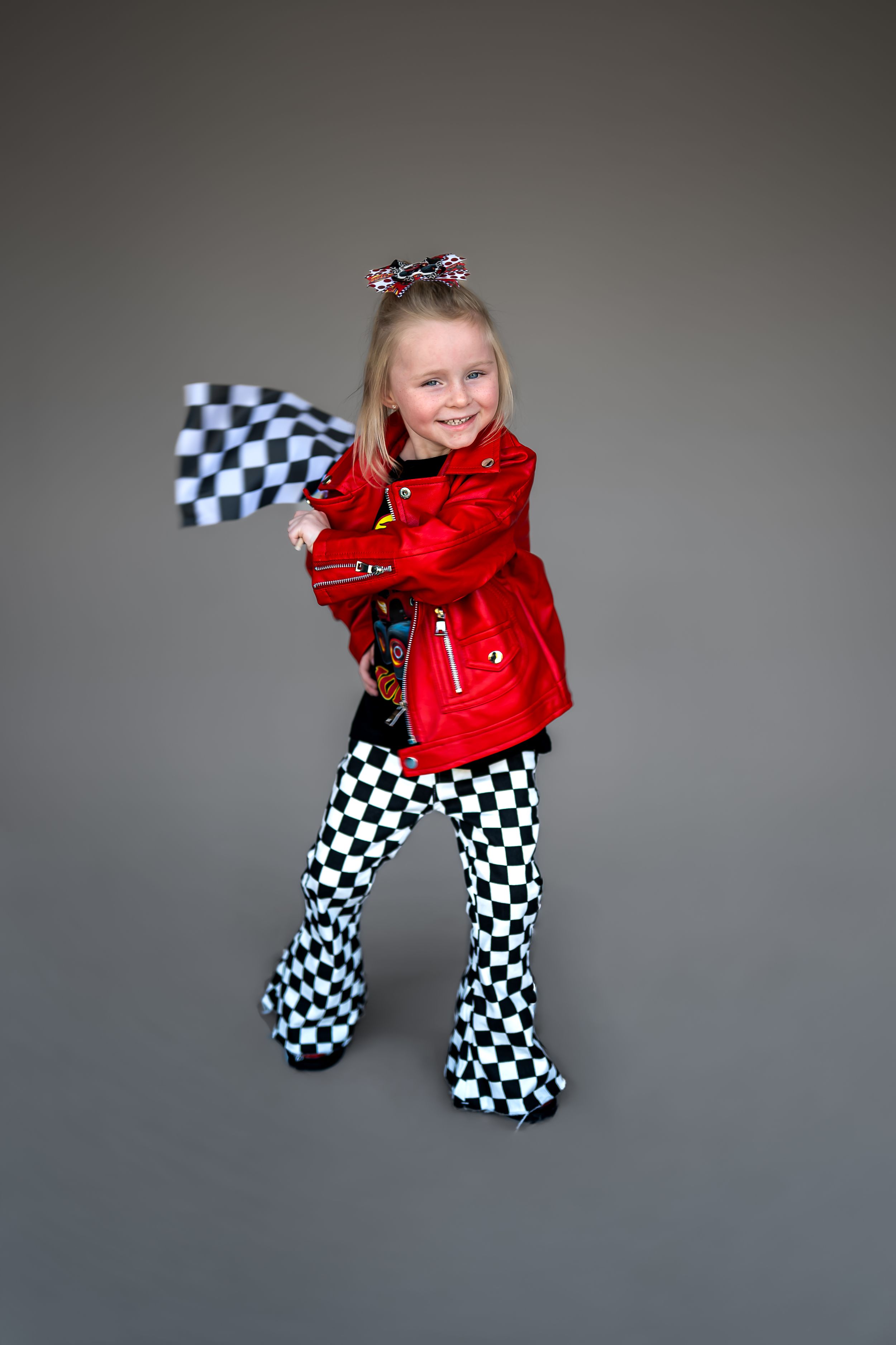 Young girl in red leather jacket and checkered pants waves a racing flag, smiling confidently against gray background.