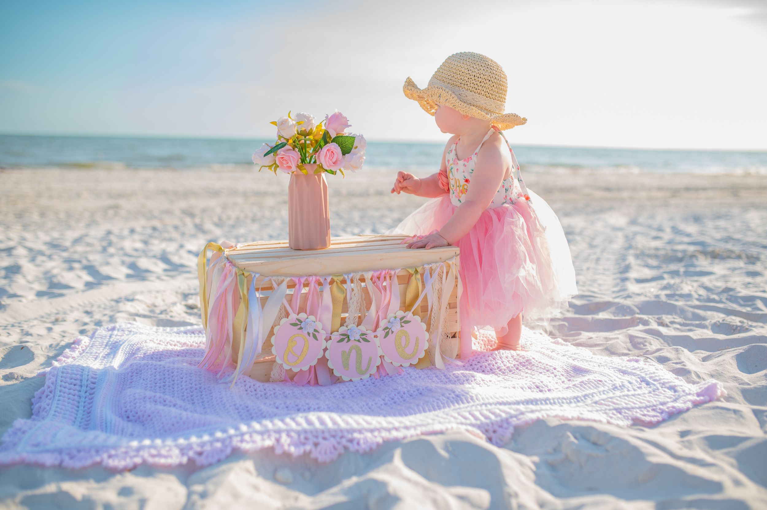 Pink tea party setup on beach blanket with flowers and ocean view.