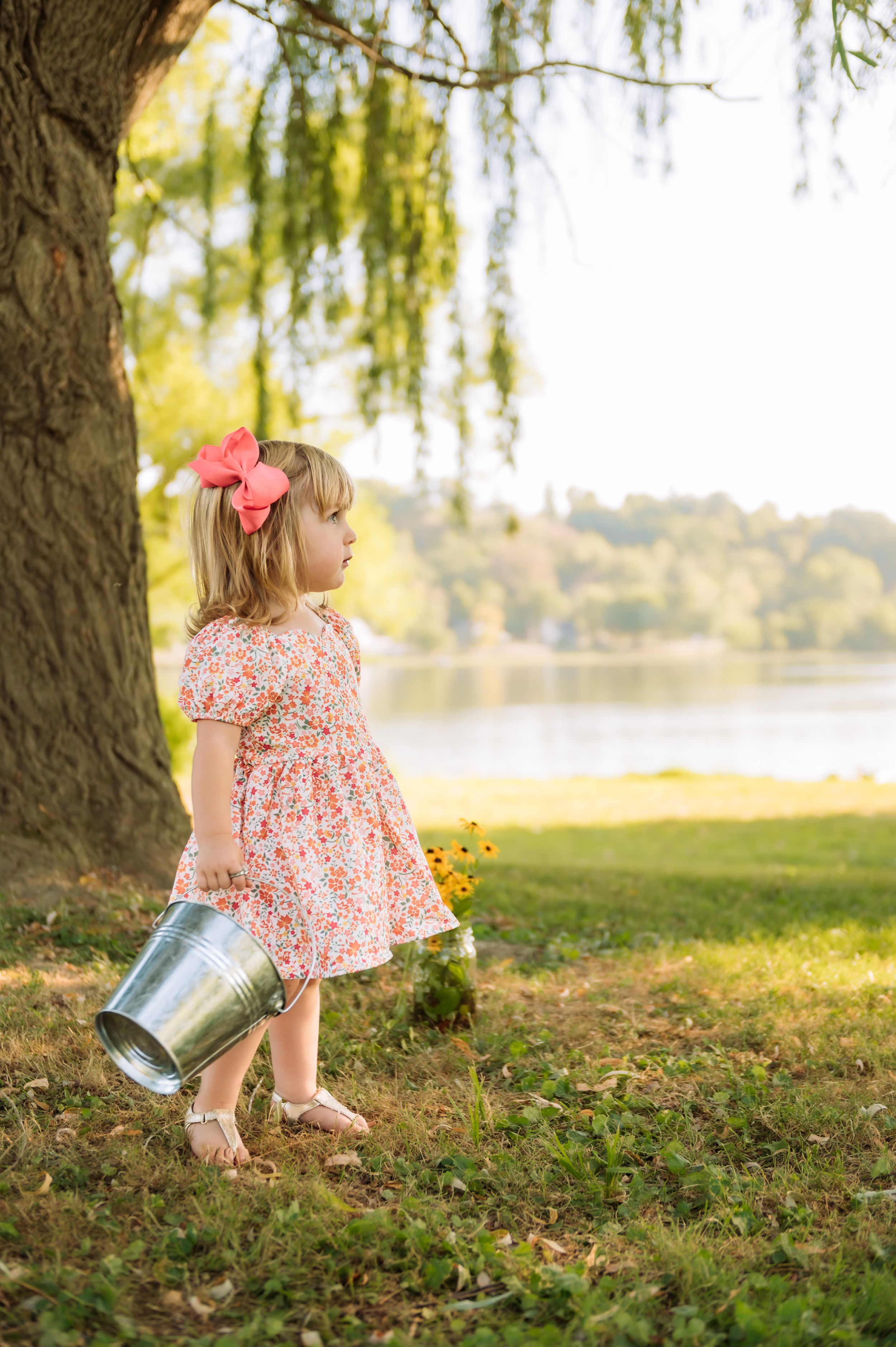 Small child in floral dress carrying metal bucket near willow tree by lake.