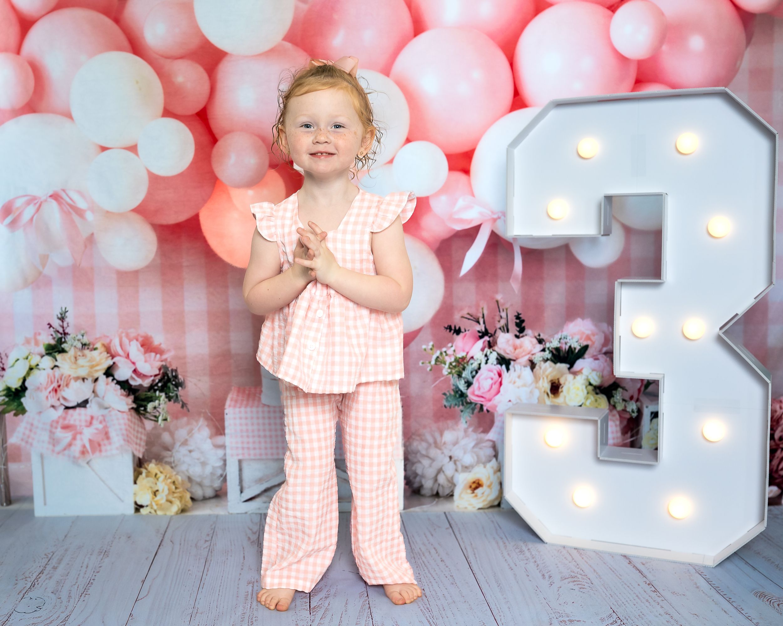 Toddler girl in pink gingham outfit poses by illuminated number 3 with pink balloons and flowers for birthday photo.