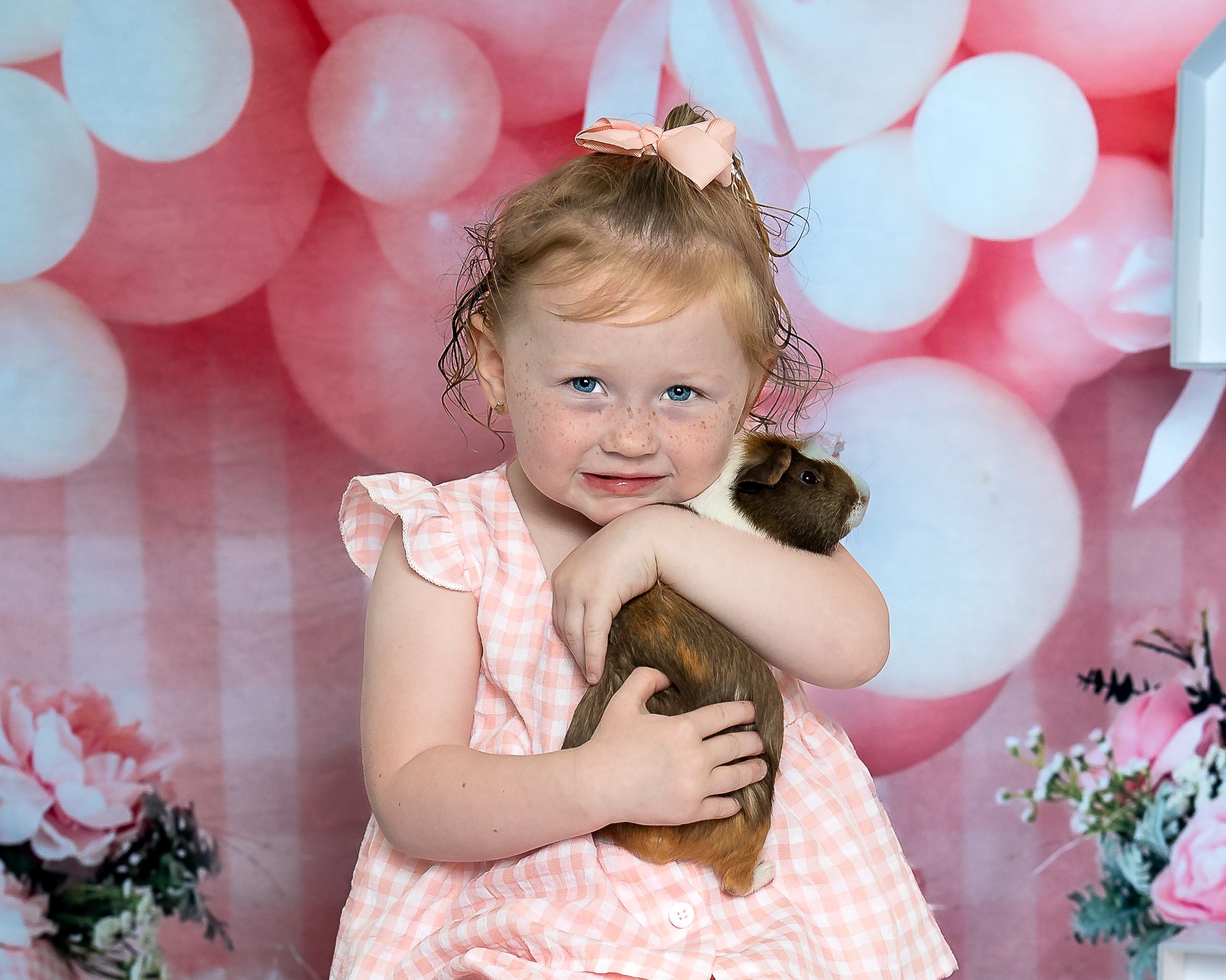 Young girl in pink dress hugging a guinea pig, smiling in front of pink and white balloon backdrop.