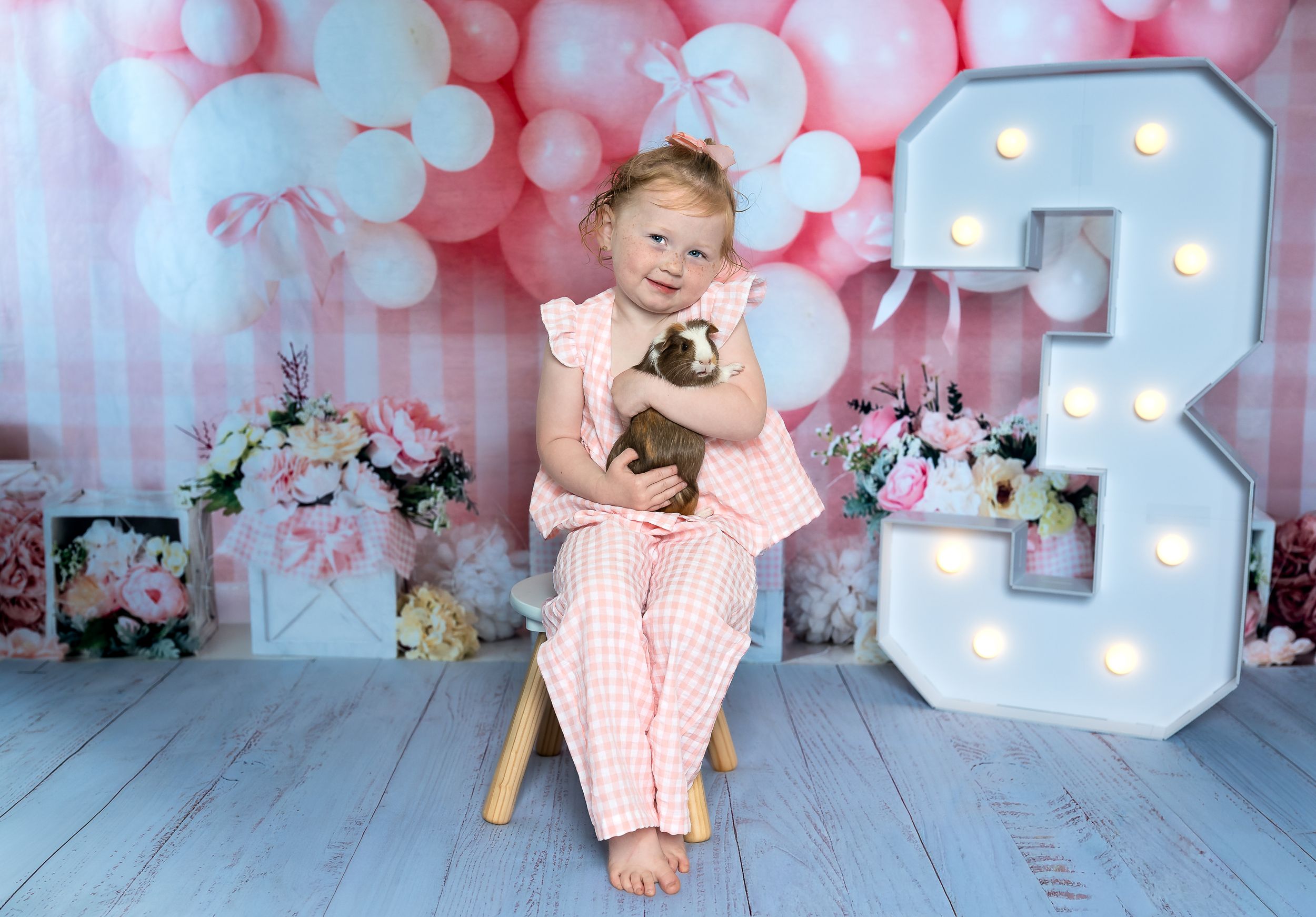 A smiling young girl in a pink gingham outfit holds a guinea pig at a festive third birthday photo shoot.