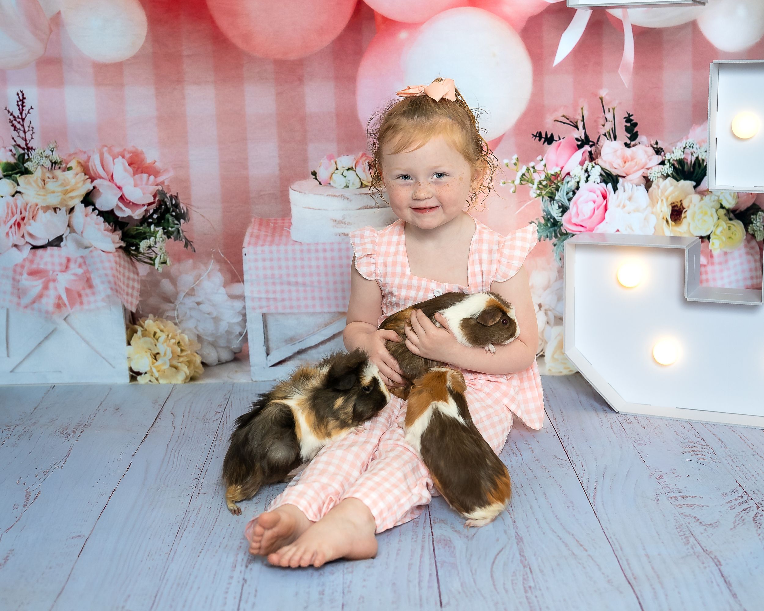 A happy toddler in a pink gingham dress sits on the floor cuddling multiple guinea pigs at a birthday shoot.