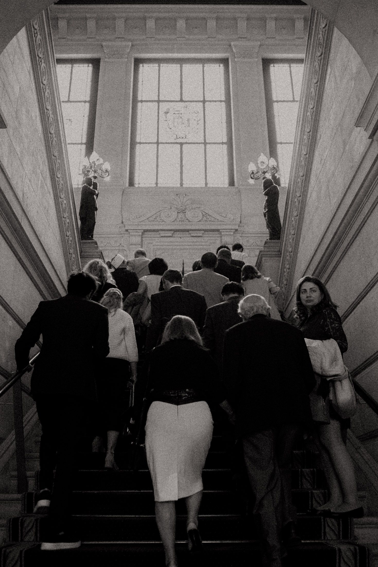 Crowd ascending elegant marble staircase in formal attire at historic building.