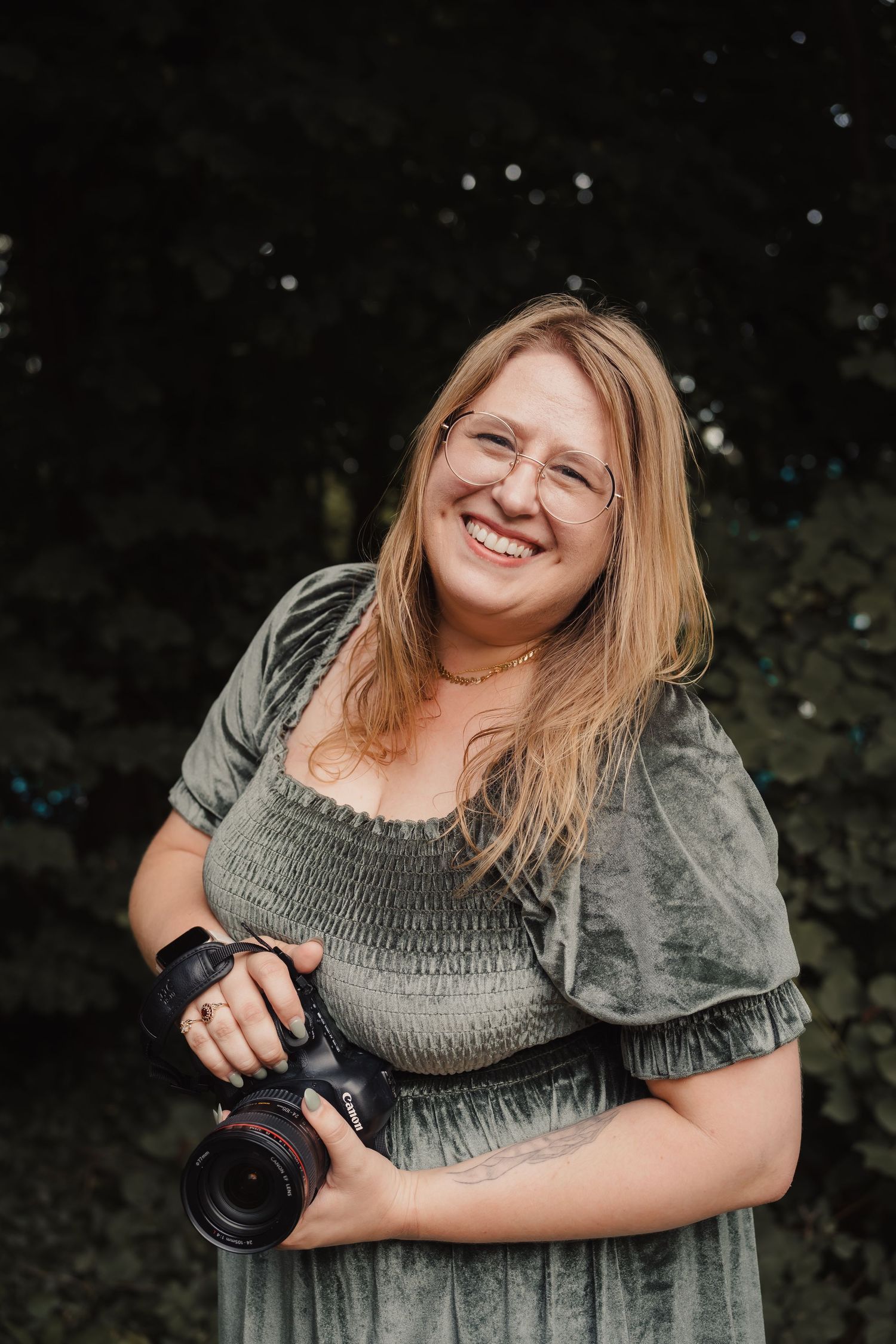 Photographer in sage green dress holding camera and smiling outdoors.