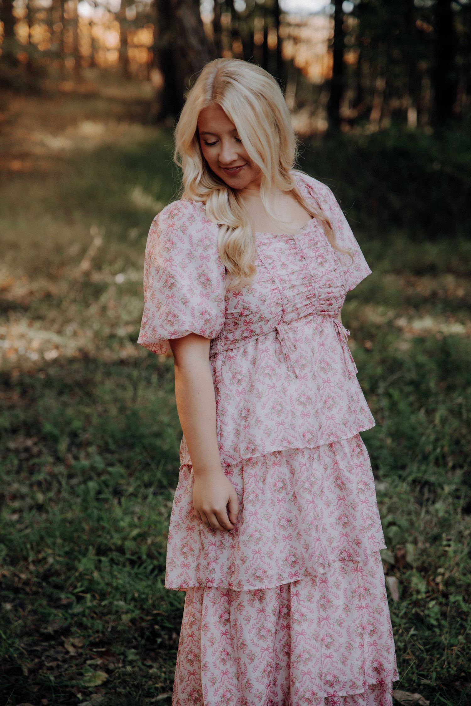 Person wearing a pink floral tiered dress poses in a wooded setting during golden hour.