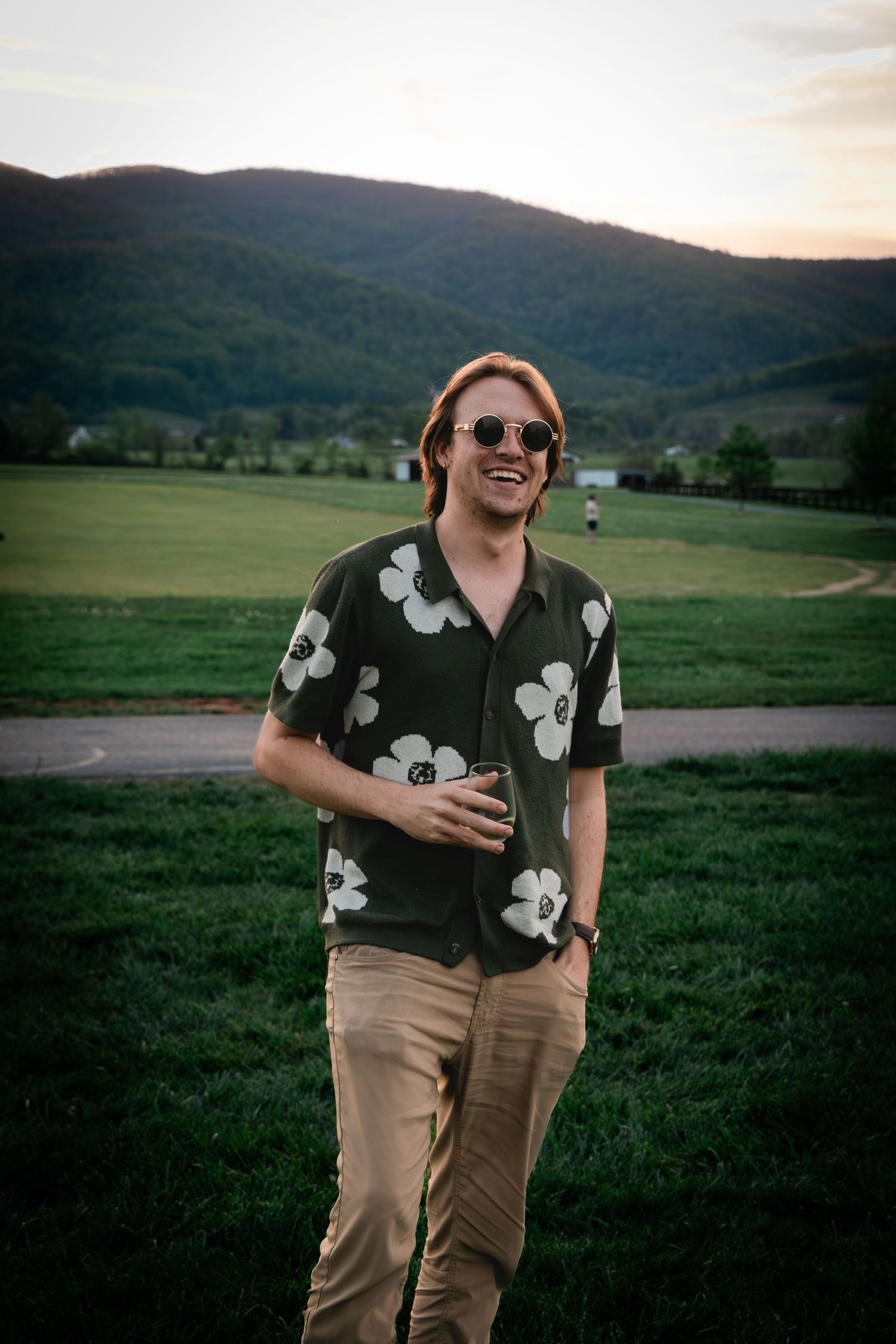 Smiling man in floral shirt and sunglasses standing in green field with mountain backdrop at sunset.