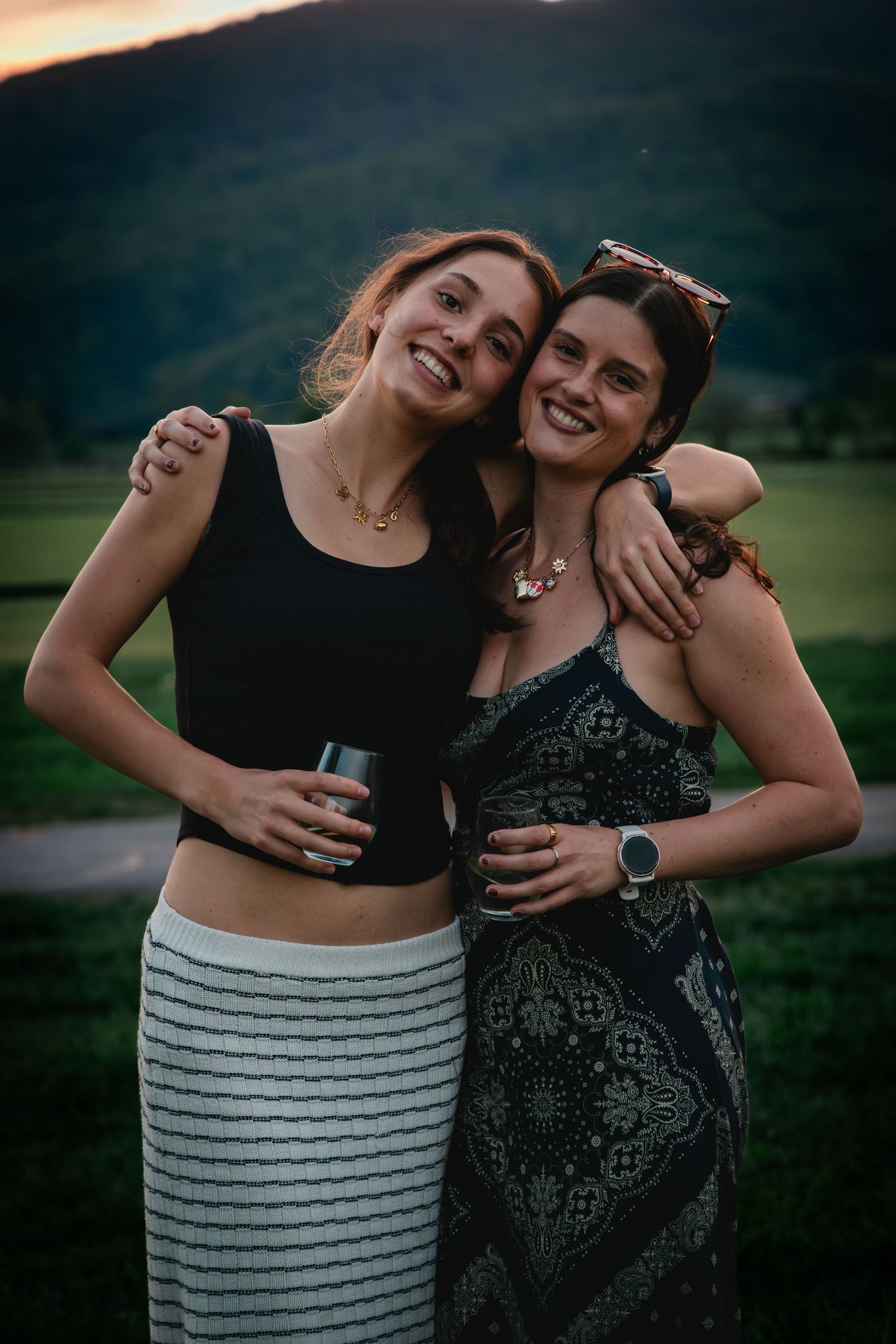 Two smiling young women hugging outdoors at sunset, holding drinks with green hills in the background.