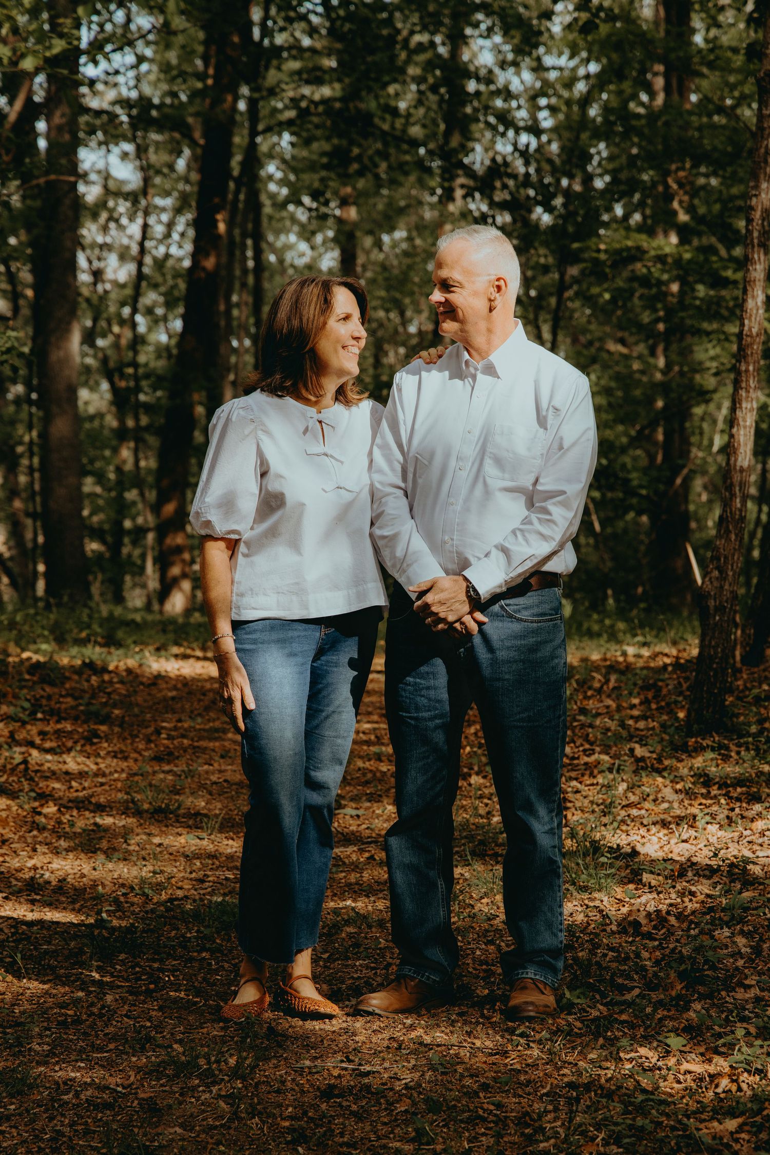 A middle-aged couple holds hands and smiles at each other in a sunlit forest wearing jeans and white tops.