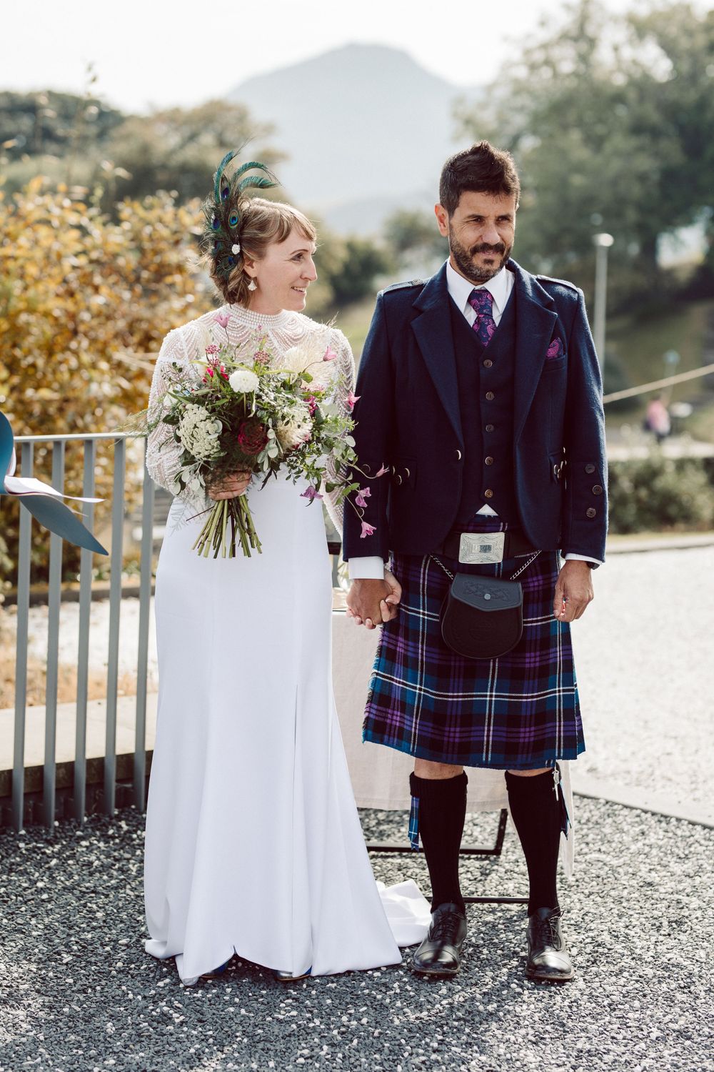 Wedding couple in traditional Scottish attire with white dress and kilt posing outdoors.