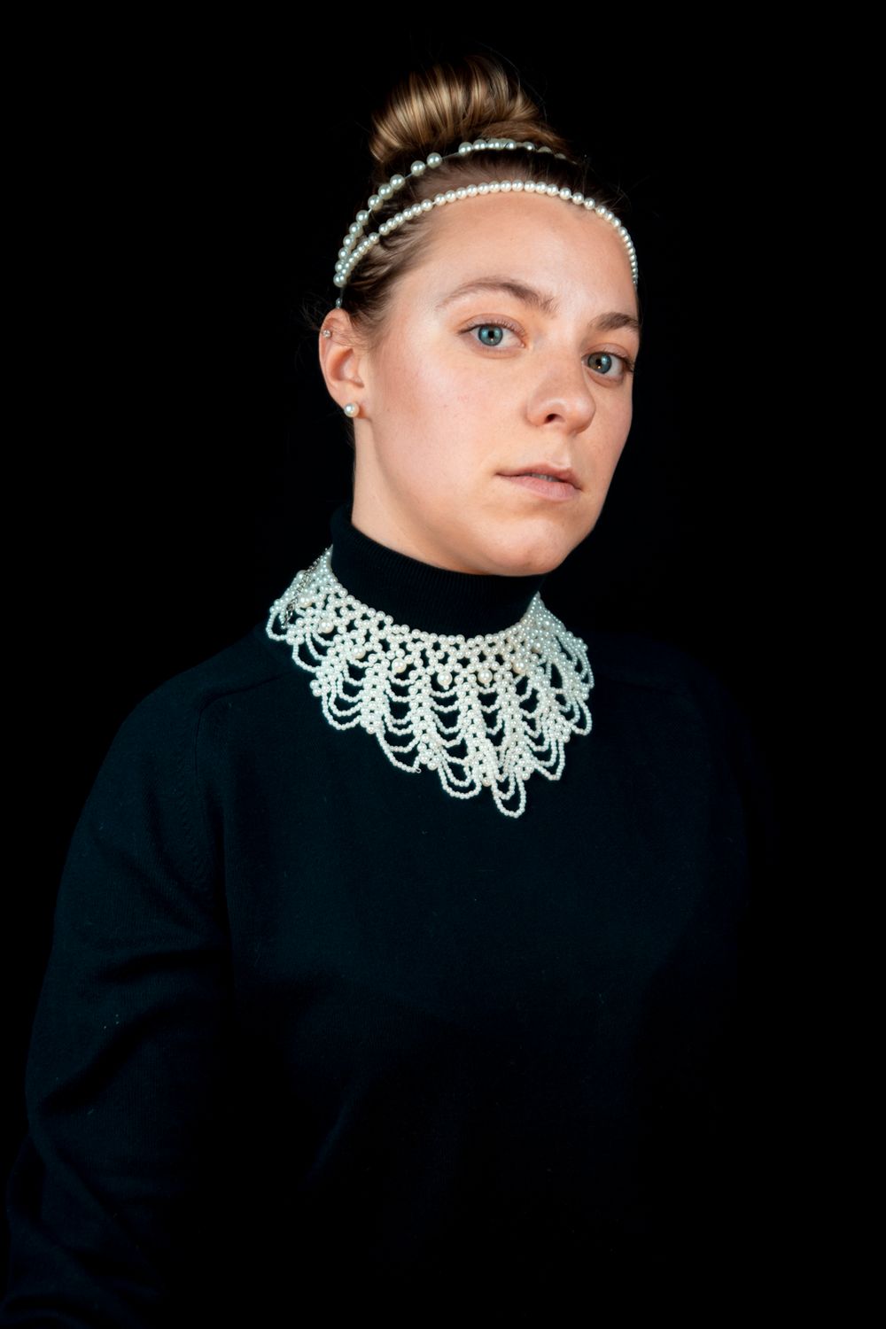 A model wearing all black with a pearl necklace, hairpiece, earrings, and ring with their head tilted.