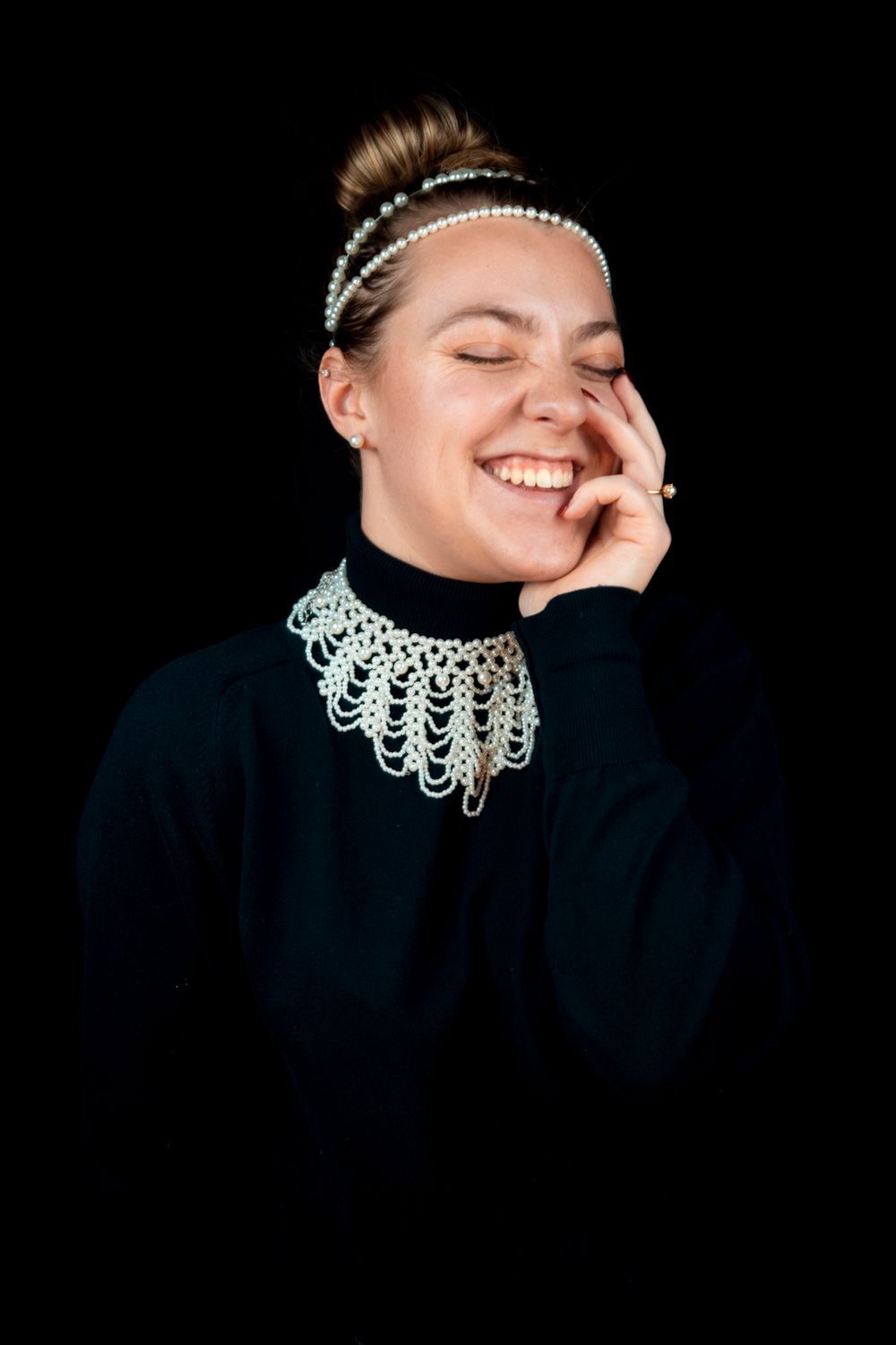 A model wearing all black with a pearl necklace, hairpiece, ring and smiling with their eyes closed.