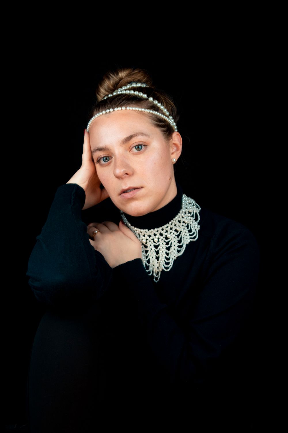 A model wearing all black with a pearl necklace, hairpiece, earrings, and ring with their hands on their shoulder.