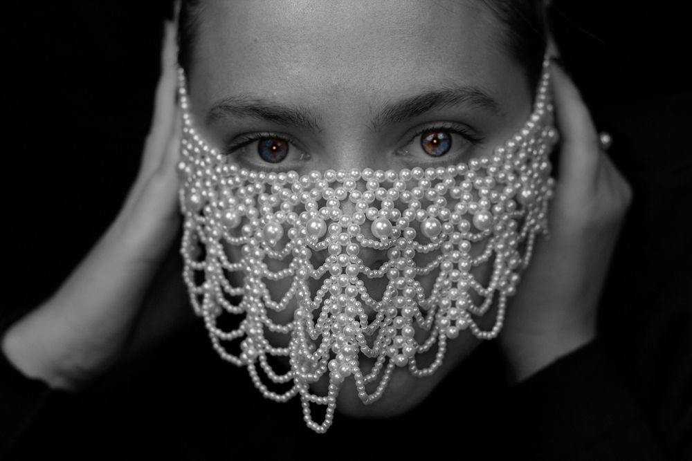 A model wearing all black with a pearl necklace covering their face looking at the camera.