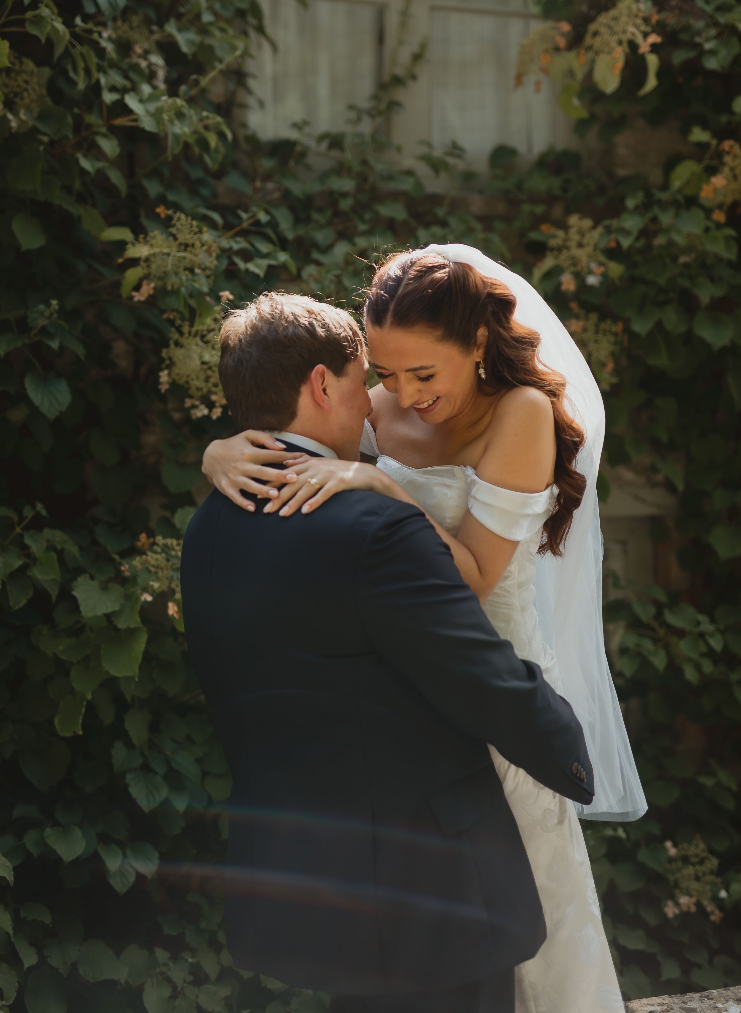 A romantic wedding moment captured outdoors with a couple embracing among lush green foliage on a sunny day.