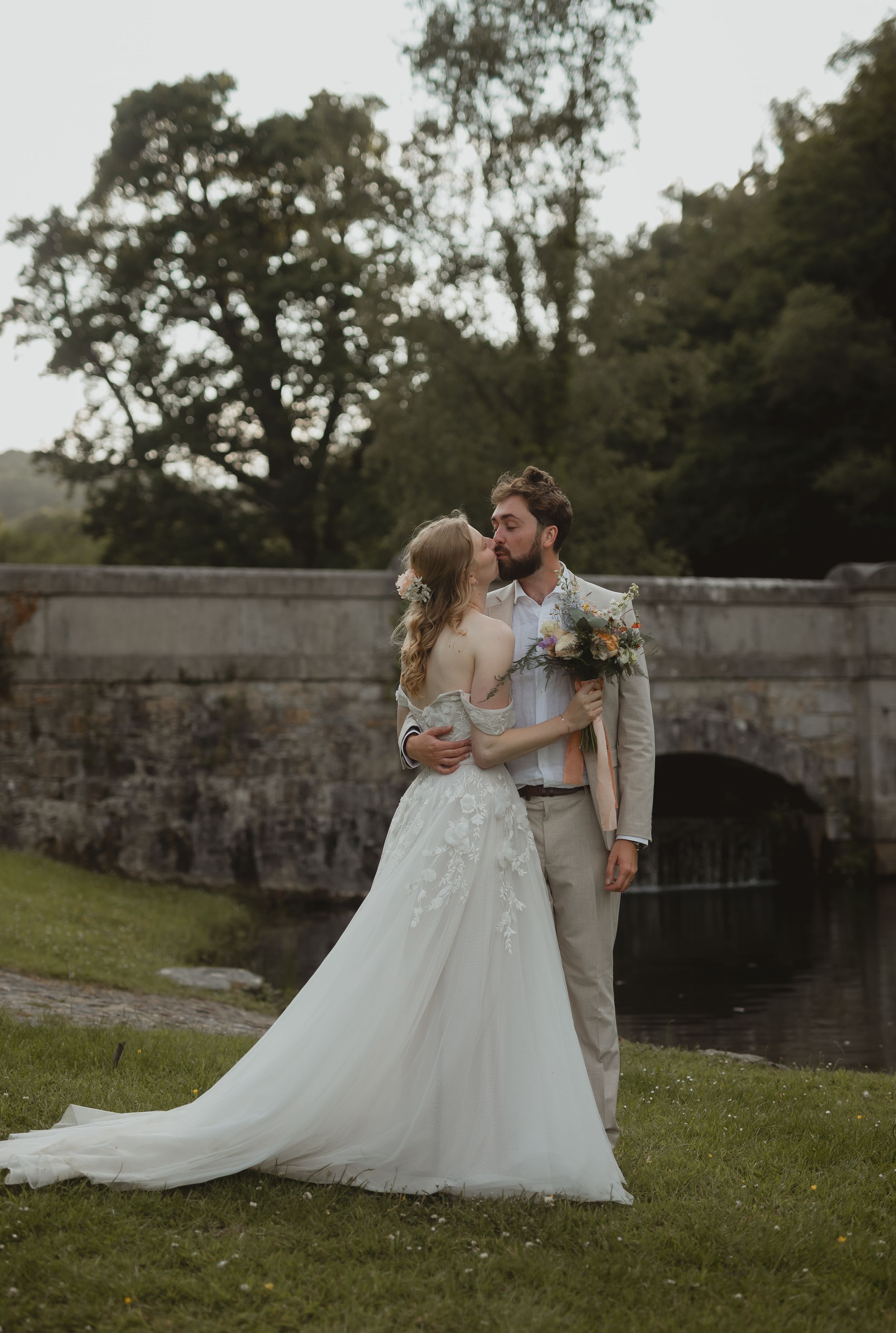Bride in flowing white gown and groom in tan suit share romantic moments by stone wall and pond during outdoor wedding shoot.