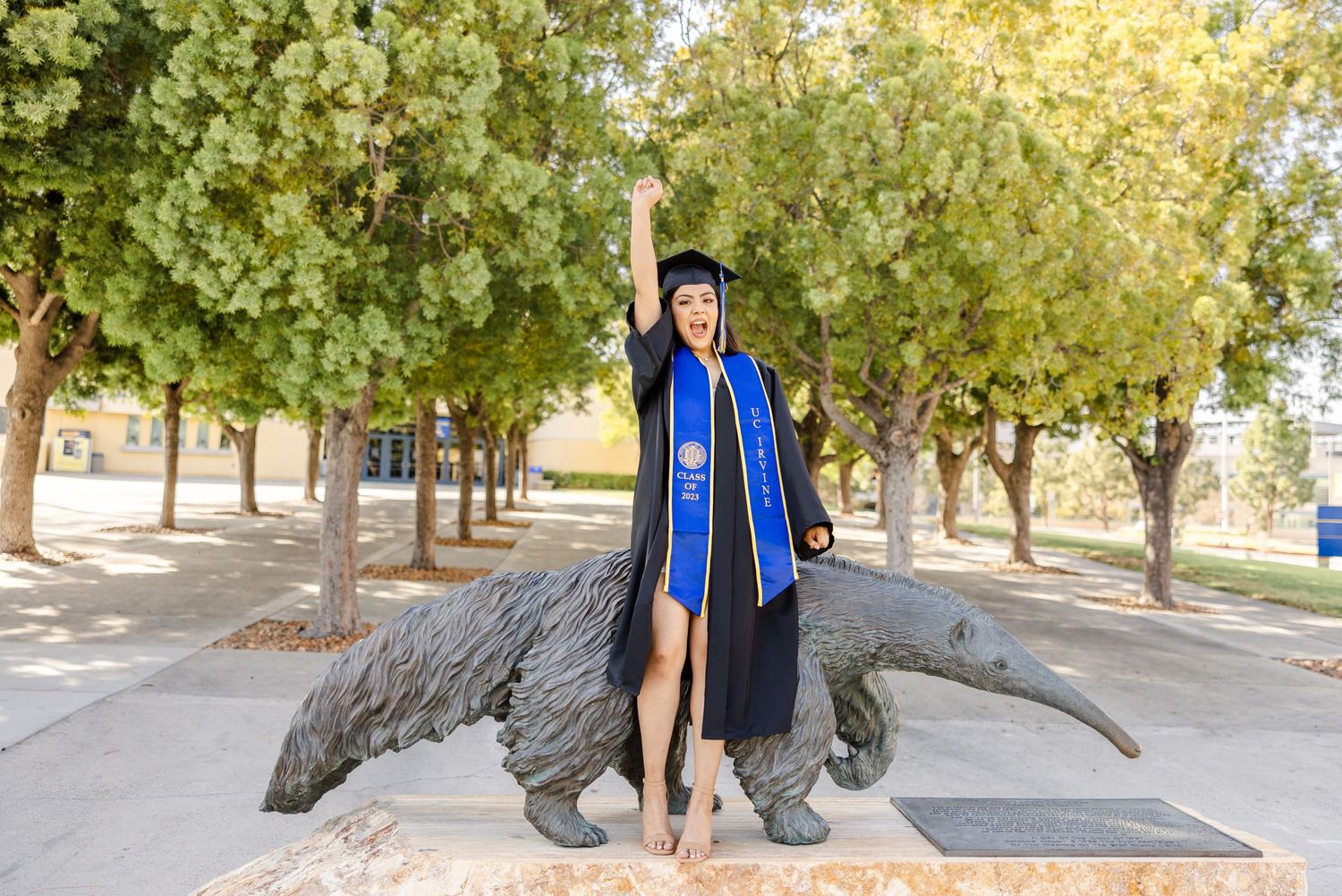 Graduate in blue cap and gown poses for celebratory photos in a sunny outdoor setting.