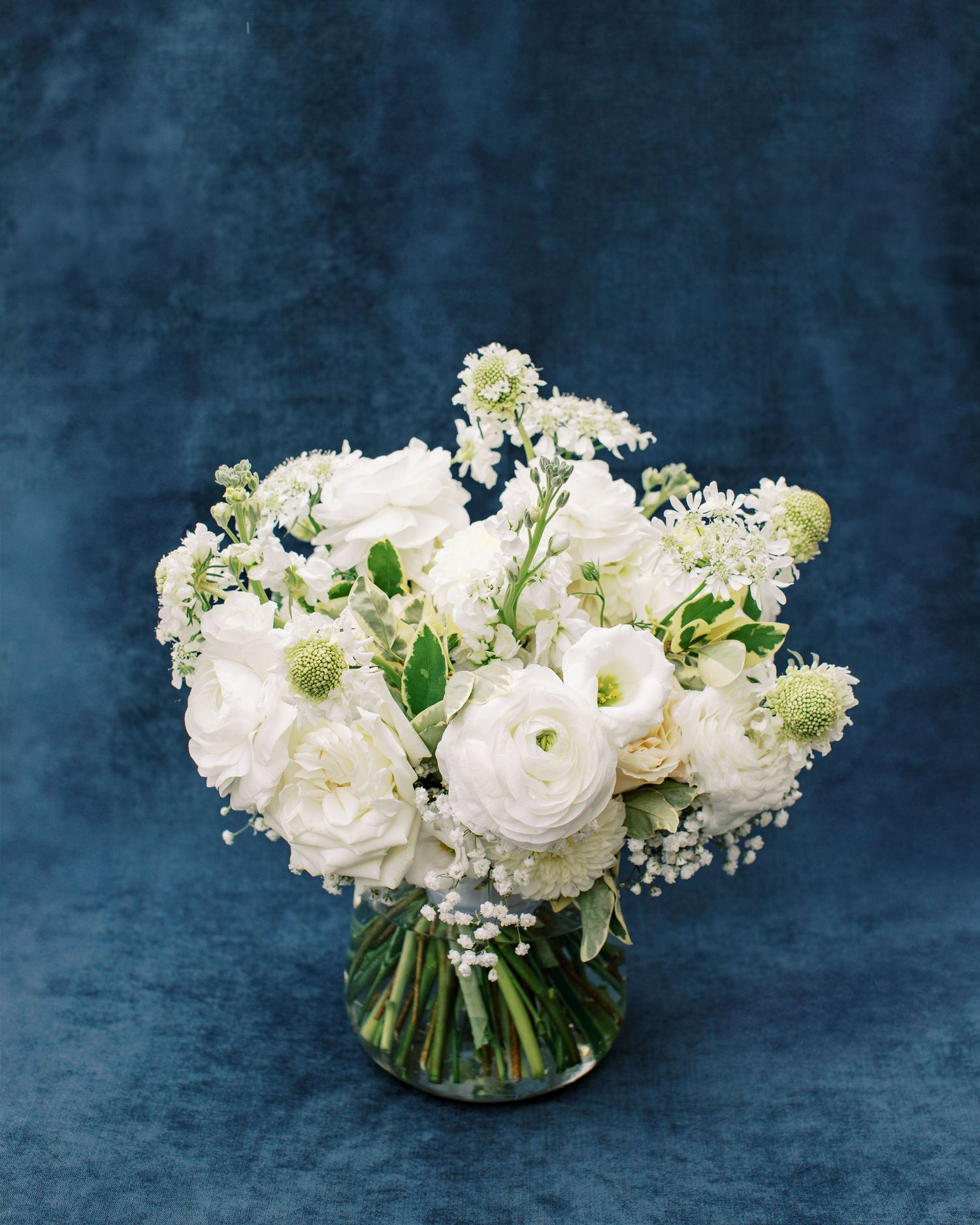 Elegant white floral centerpiece with ranunculus and baby's breath in a glass vase against blue backdrop.
