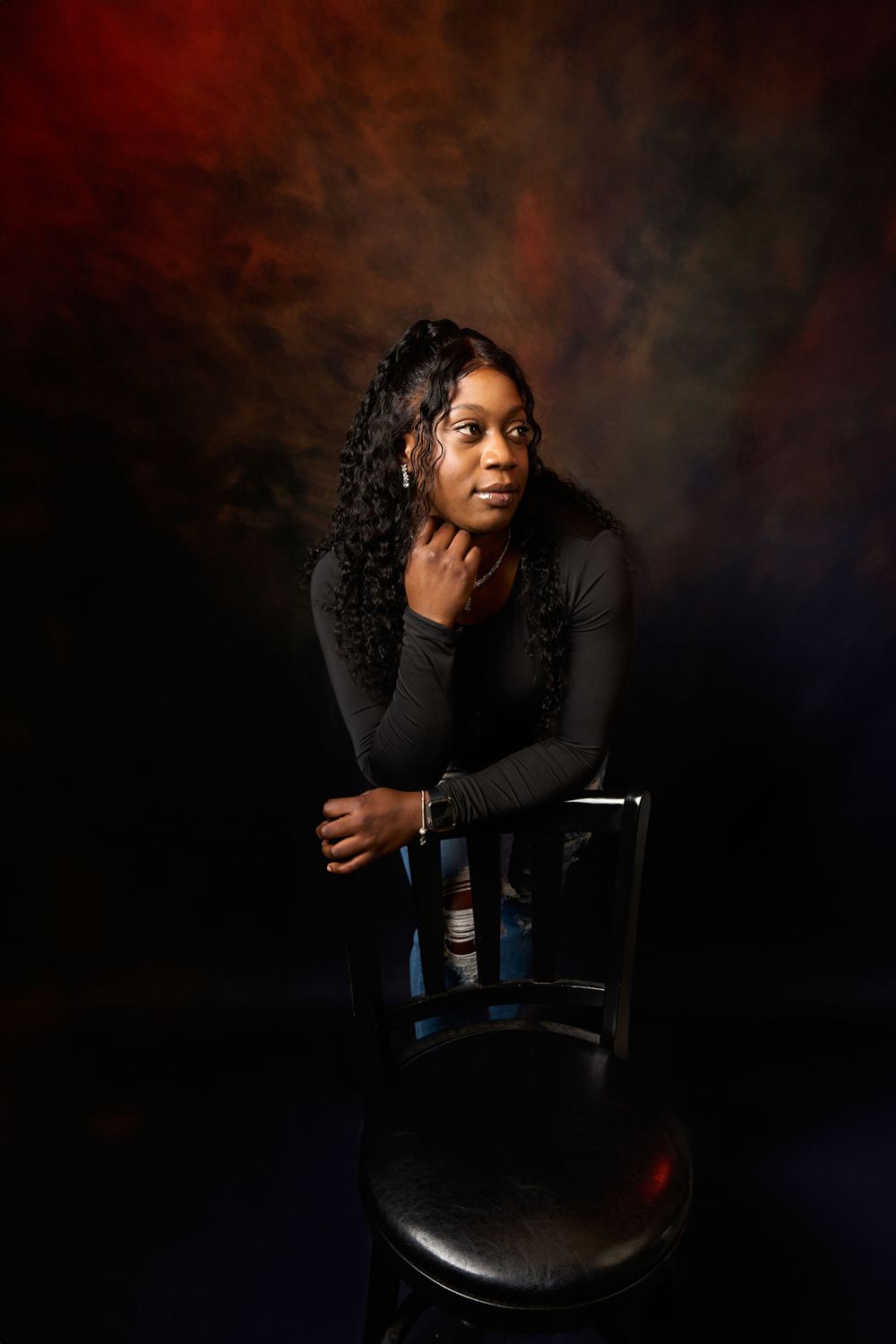 Studio portrait in dark setting showing model in black attire posing on chair against dramatic background.