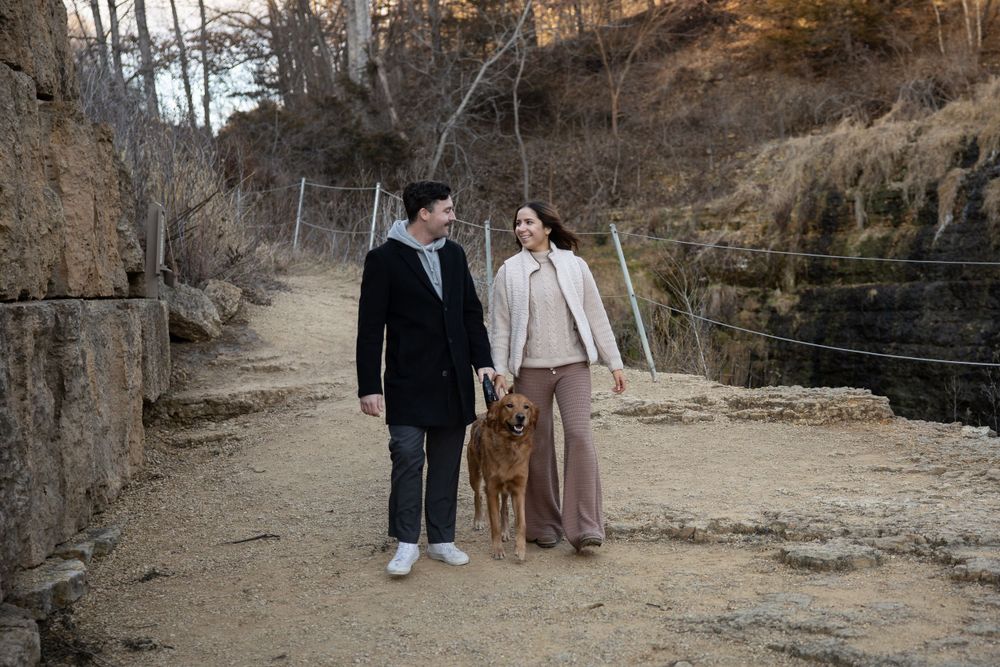 A couple walks with their dog along a trail with cliffs and waterfalls in the background.