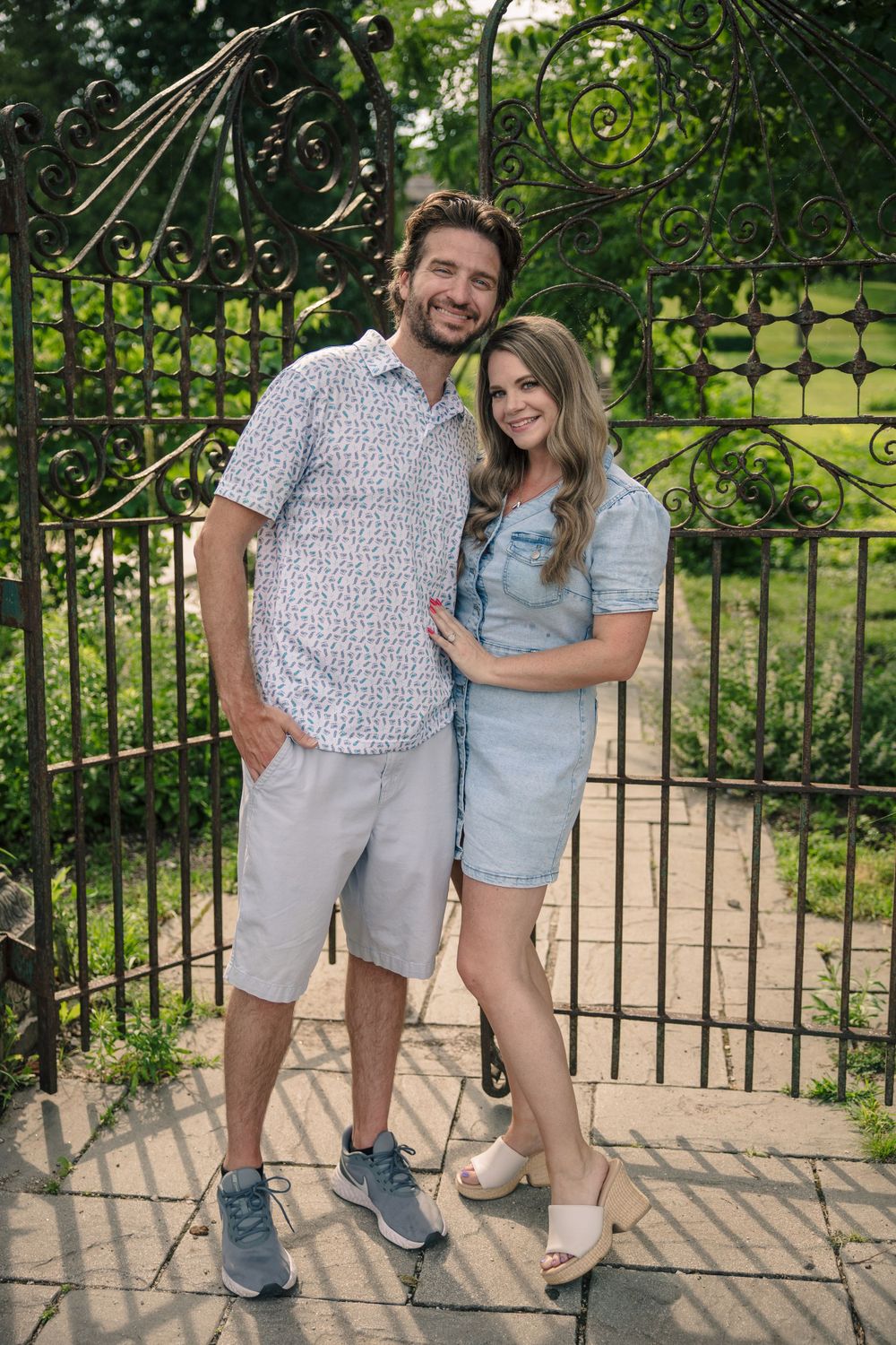 A couple poses together in casual summer outfits by a decorative iron gate in a garden setting.