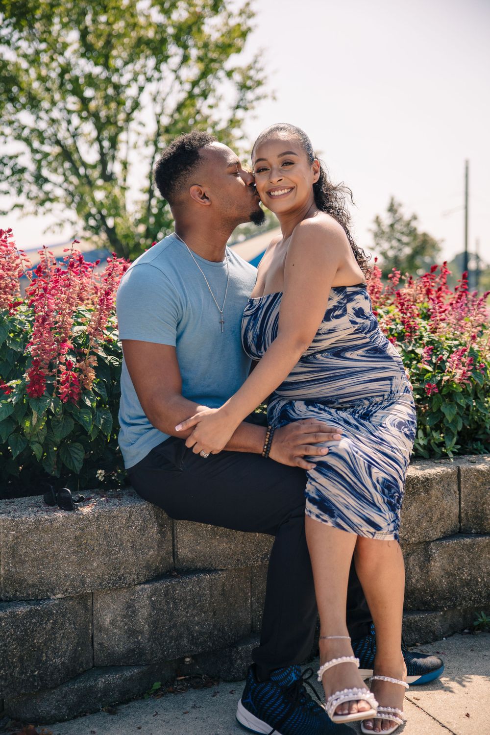 Couple sharing an intimate moment while sitting on a stone wall with pink flowers in background.