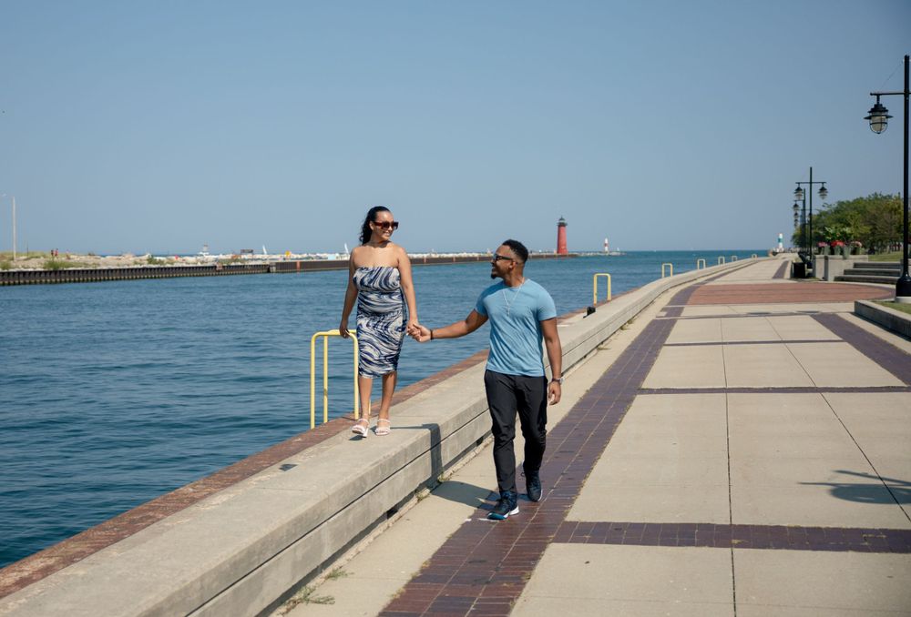 A couple takes a romantic stroll along a waterfront promenade with a lighthouse visible in the distance.