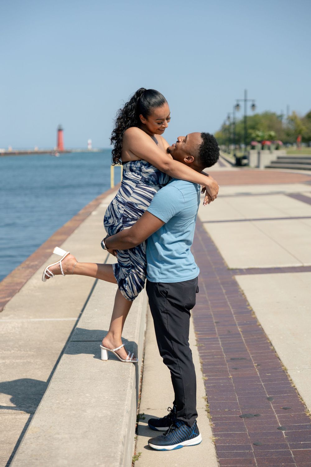 A romantic couple embraces on a waterfront promenade with a red lighthouse in the background.
