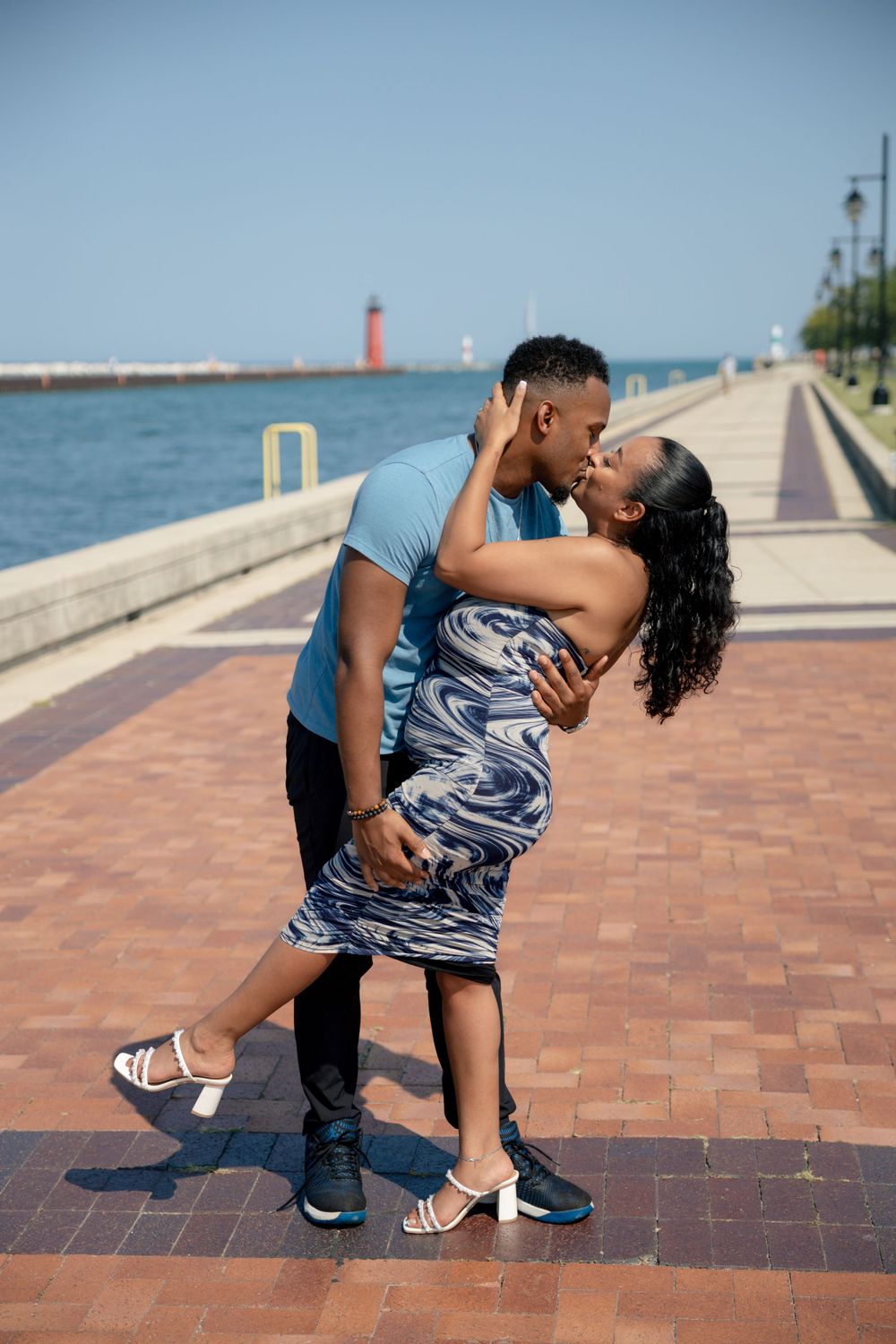A couple shares an intimate dance pose on a brick waterfront walkway with a lighthouse in view.