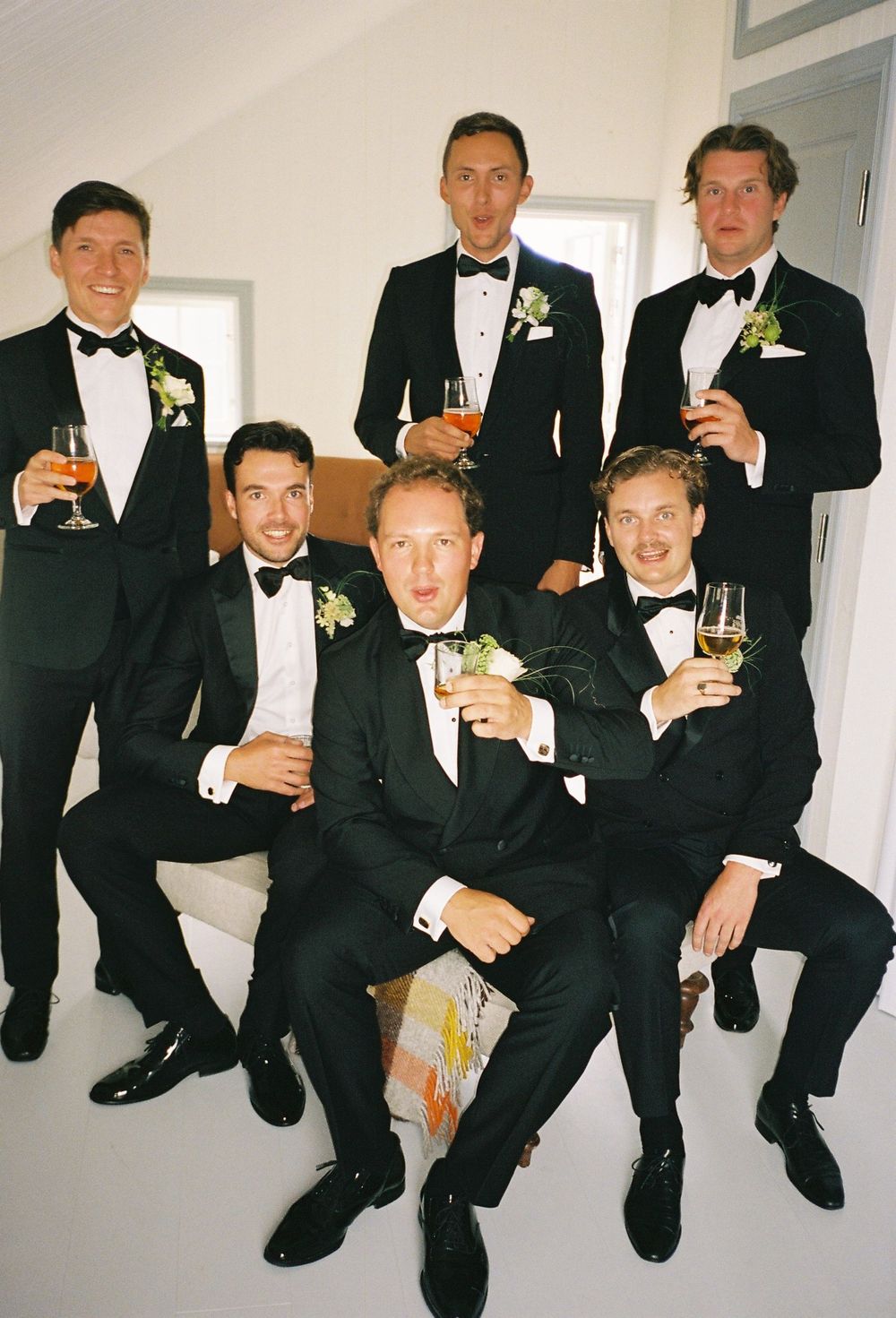 Six groomsmen in black tuxedos with boutonnieres raising glasses for a toast at a wedding celebration.