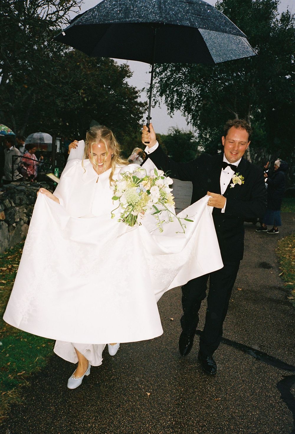 A bride and groom walk in the rain, she holds her white gown while he shields them with a black umbrella.