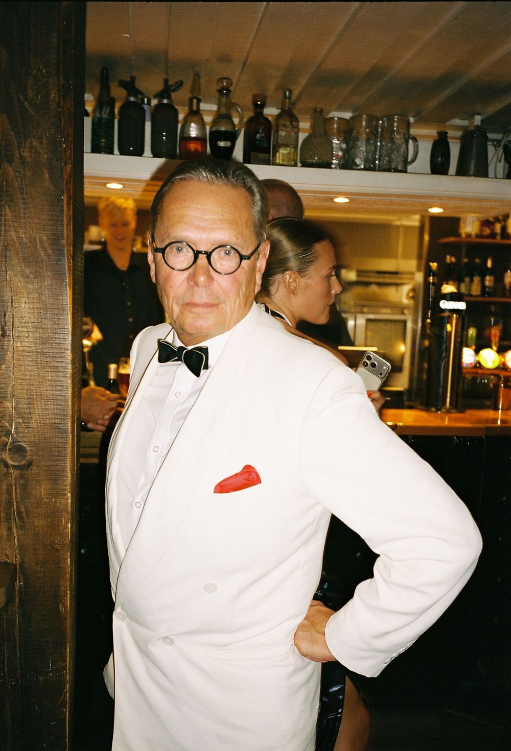 Elegant man in white tuxedo jacket with red pocket square and bow tie poses at a bar.