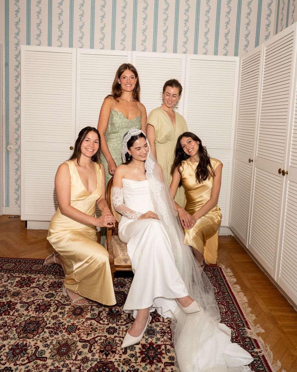 Bride in white gown with veil poses with four bridesmaids in gold and green satin dresses in elegant room.