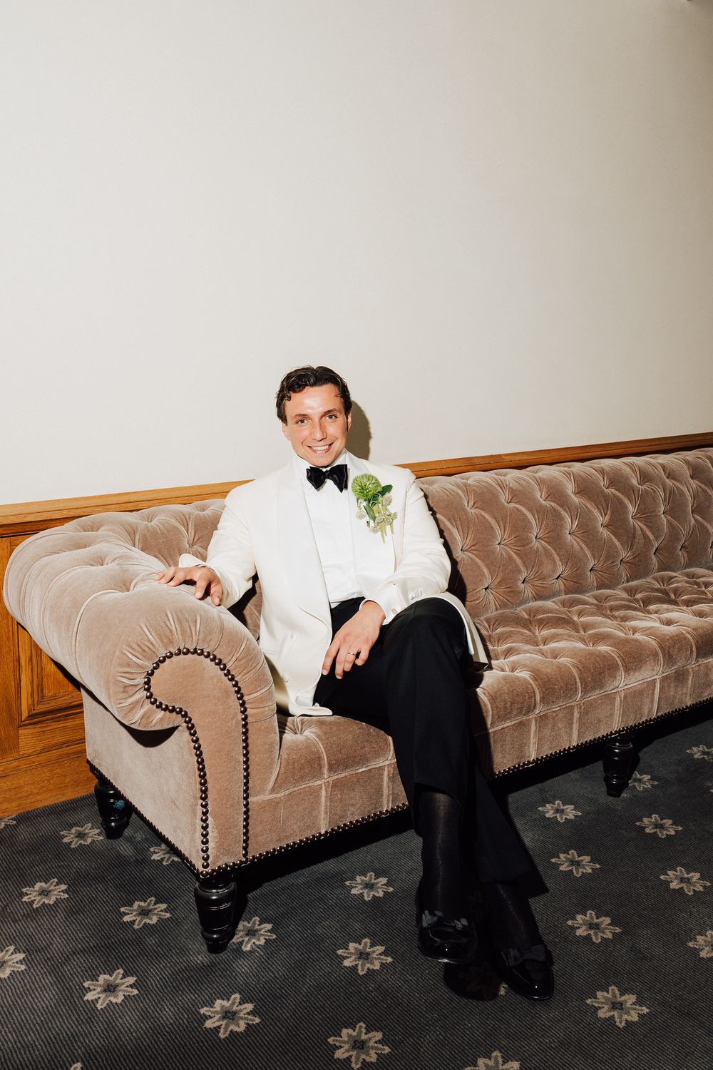 Smiling groom in white tuxedo jacket and black bow tie sits on tufted velvet sofa with green boutonniere.
