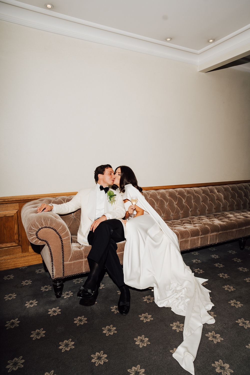 Bride and groom share a romantic kiss while seated on a vintage velvet sofa in an elegant wedding venue.