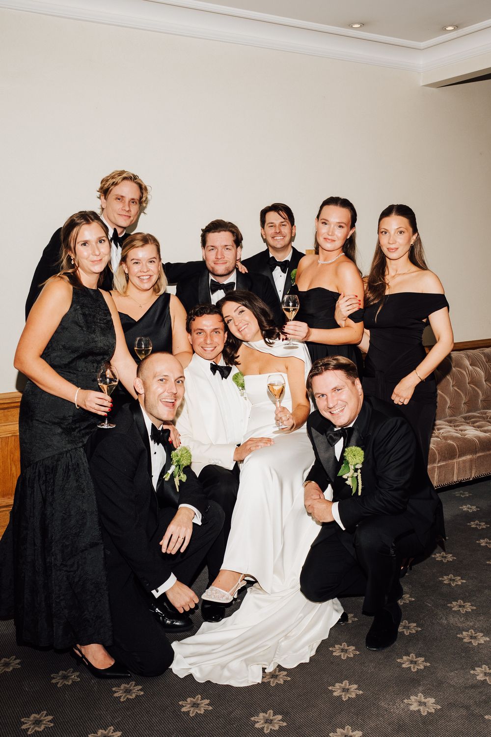 Wedding party in black formal attire poses with bride in white gown, holding champagne glasses for a group photo.