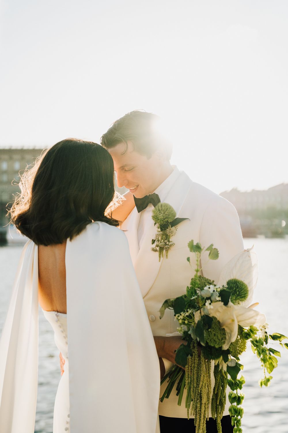 Bride and groom share a romantic moment at sunset near waterfront, groom holding elegant white and green bouquet.