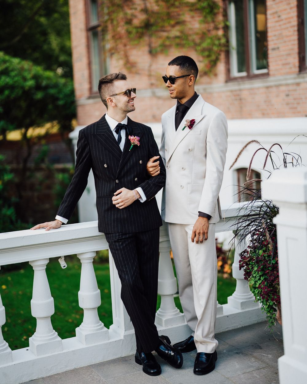 Two grooms in elegant suits posing together on balcony with boutonnières on wedding day.
