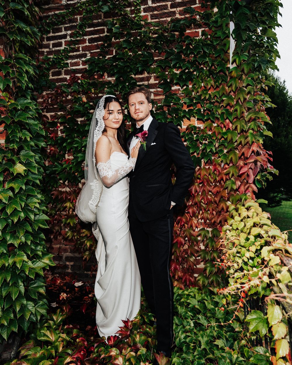 Bride and groom pose elegantly before ivy-covered brick wall, she in lace gown with veil, he in classic black tuxedo.