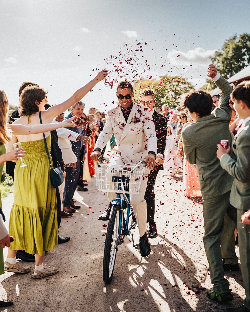 Groom in white suit rides bicycle through colorful confetti thrown by cheering wedding guests.