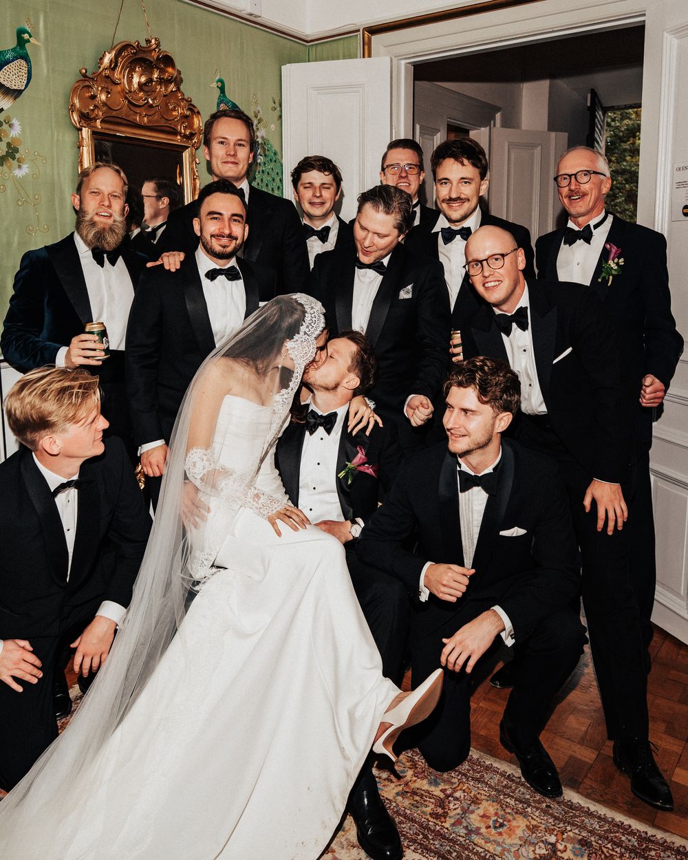 Bride and groom share a kiss surrounded by groomsmen in black tuxedos during a wedding party photo indoors.