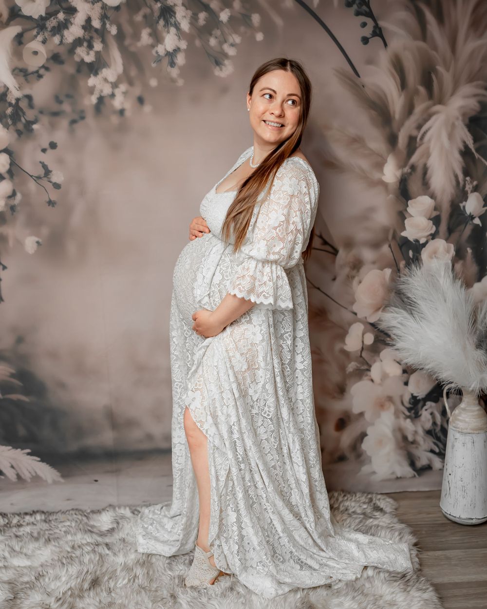 Full length maternity portrait in flowing white lace gown with floral backdrop.