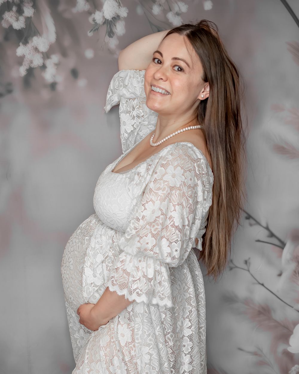 Intimate maternity portrait in white lace dress against soft floral background.
