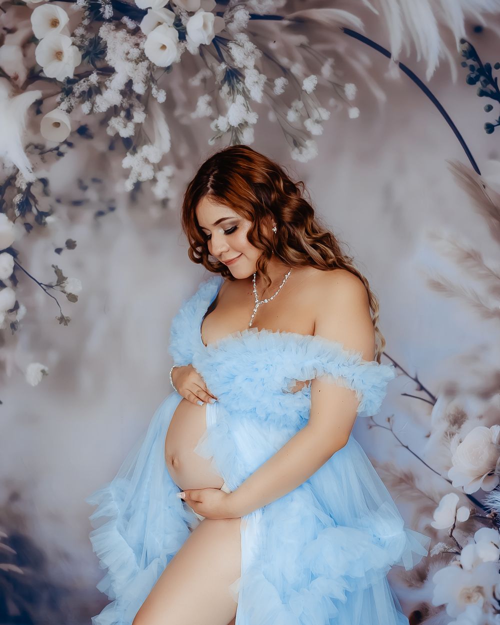 Expecting mother in flowy blue dress poses amid floral backdrop.