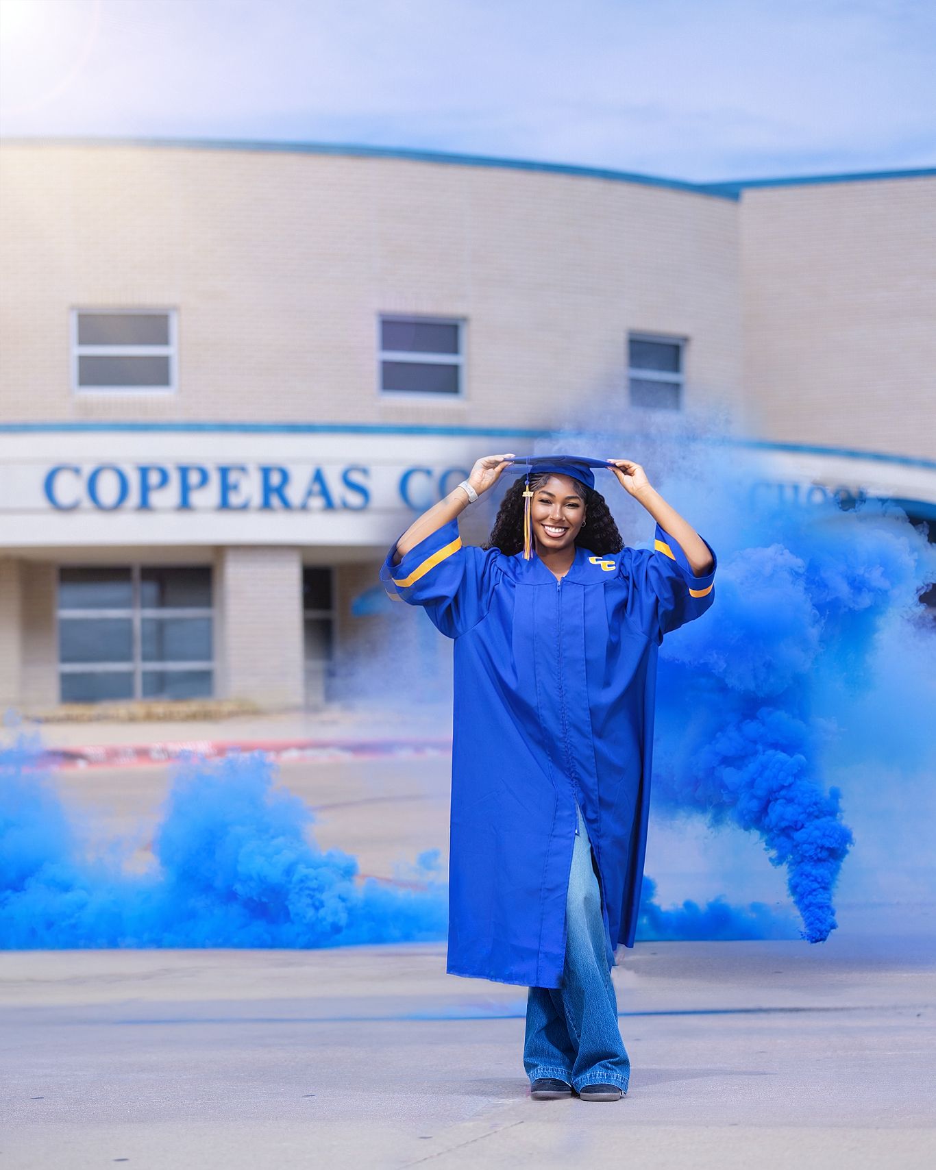 A smiling graduate in blue cap and gown poses with blue smoke bombs outside Copperas Cove High School.
