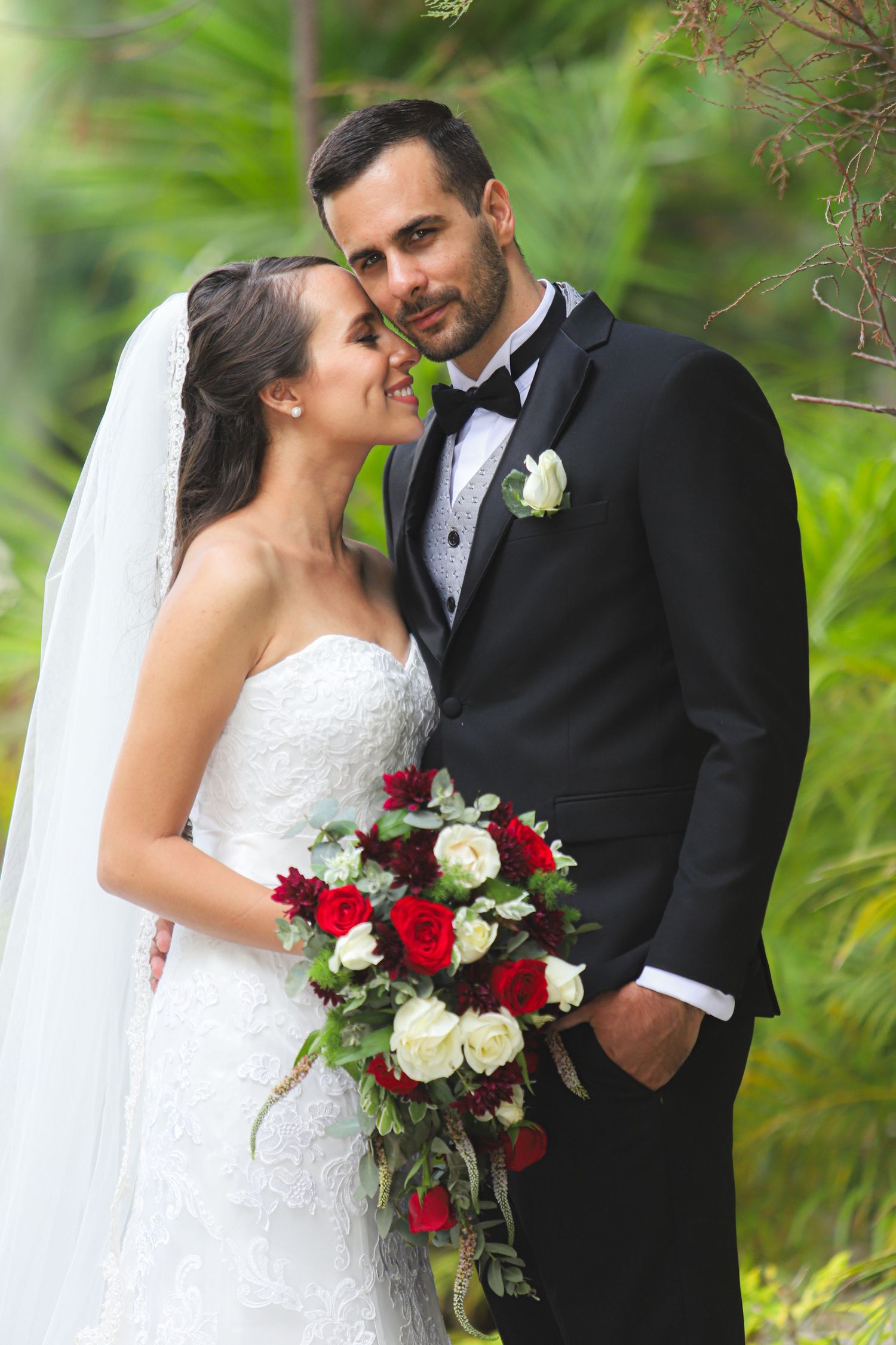 A wedding couple in formal attire stand close together outdoors with a red and white bouquet against a green background.